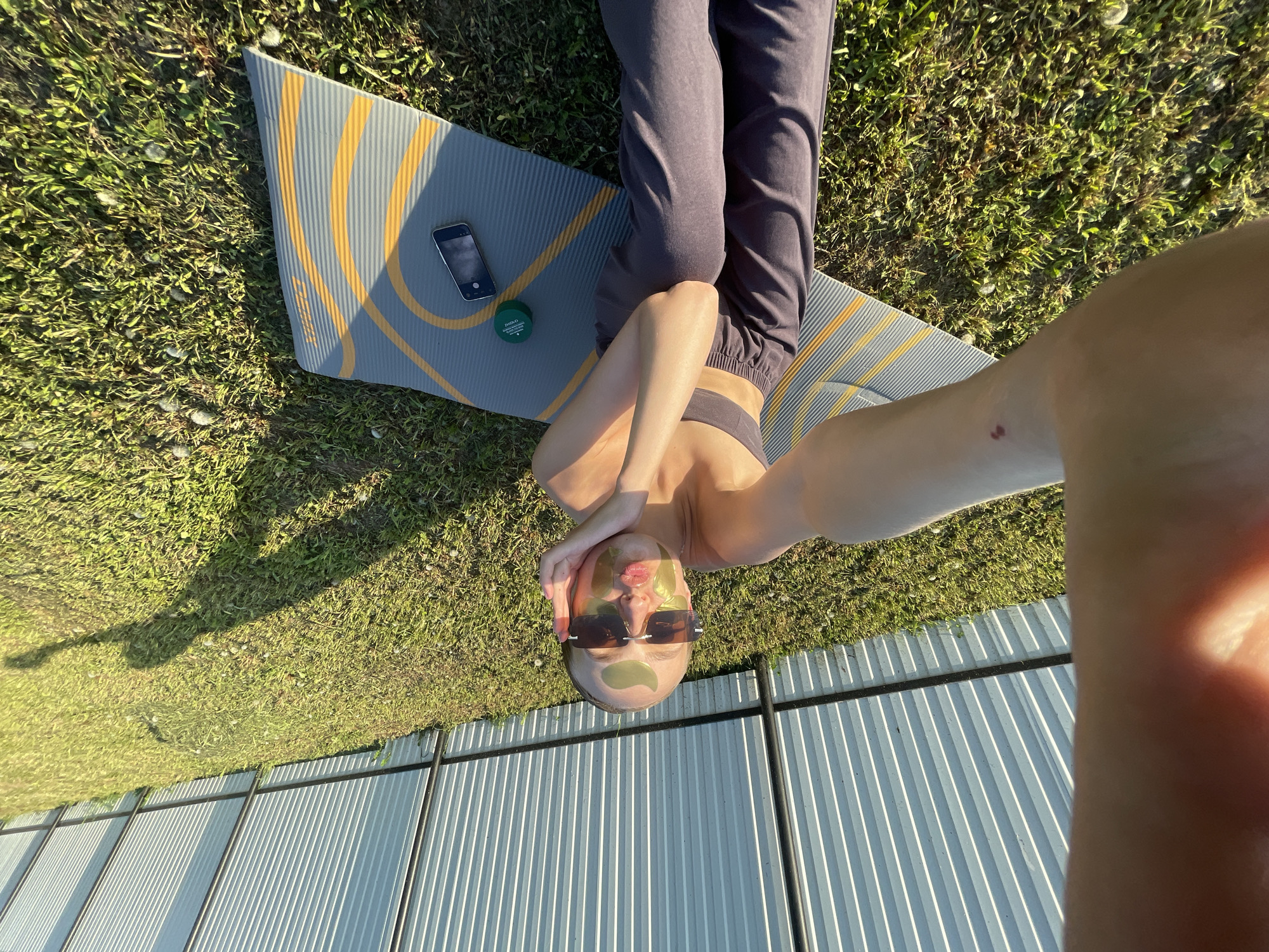 The image is a selfie taken from an upward angle. A person is lying on a gray yoga mat with yellow stripes, which is on green grass. The person is wearing dark athletic pants, a dark sports bra, and sunglasses. Green patches are visible under their eyes. A green container and a smartphone are also on the yoga mat. The edge of a corrugated metal fence or building is visible in the foreground. There is an indication that the location is in Подшивалово, Russia.
