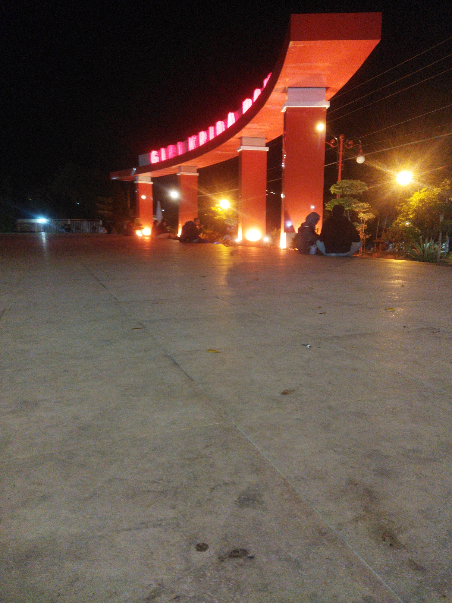 The image depicts a nighttime scene at the Giri Krida Bakti tourism area in Wonogiri, Indonesia. A large, illuminated archway with "GIRI KRIDA BAKTI" in red neon lettering spans across the frame, supported by substantial red pillars. Several people are seated on the ground in front of the archway, illuminated by warm-toned artificial lights. The ground in the foreground is a paved, light-colored surface, showing some wear and tear. Trees and streetlights are visible in the background, contributing to the ambient illumination of the area. The overall atmosphere is calm and relaxed, suggesting a public space being enjoyed by visitors at night.