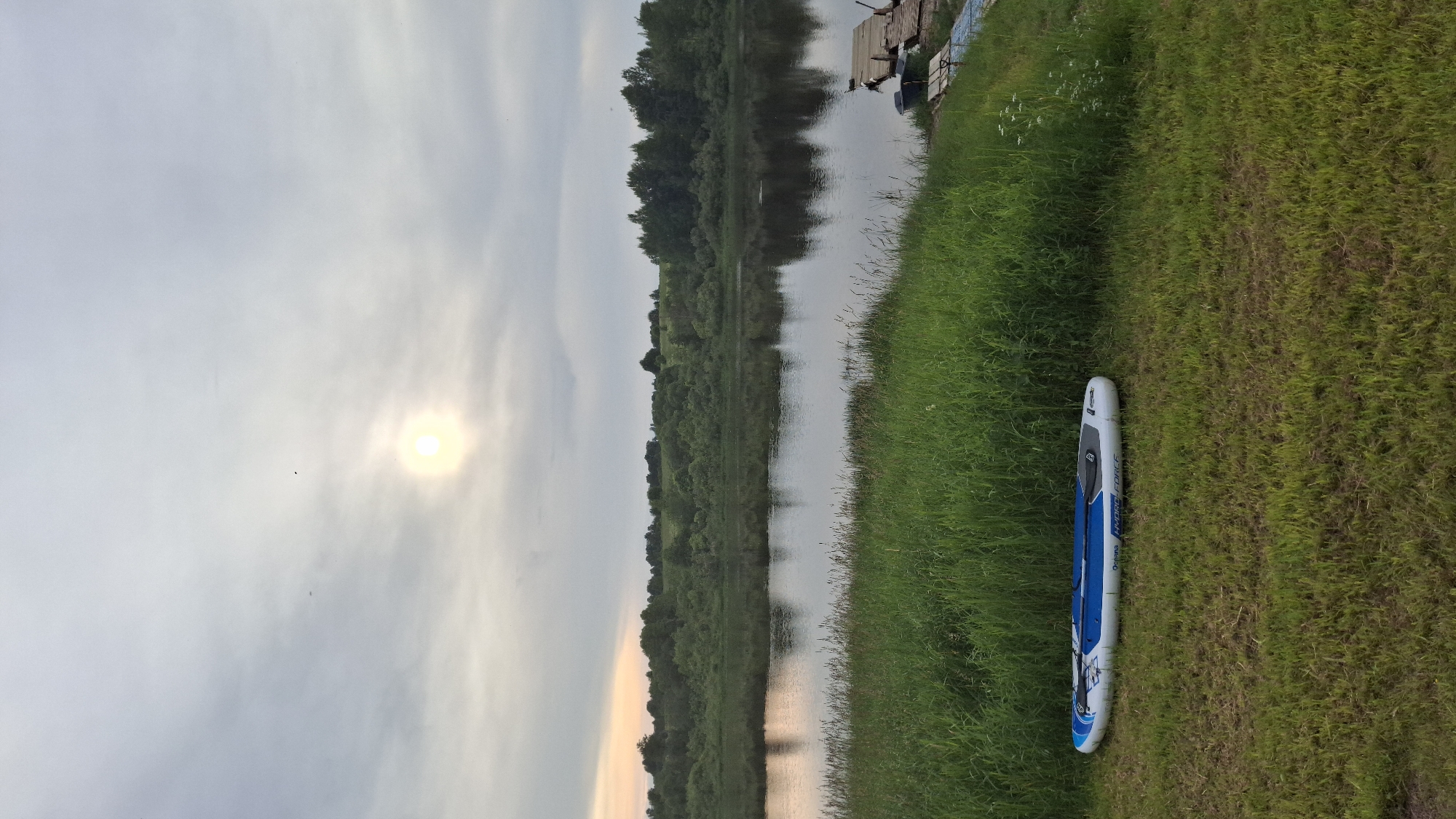 This is a landscape photograph of a riverbank with a paddleboard lying on the grass. The sun is low in the sky, casting a soft glow through a hazy cloud cover, suggesting it is either early morning or late afternoon. The river stretches into the distance, lined with lush green trees on the opposite bank, their reflections visible on the calm water's surface. A wooden dock is partially visible on the right side of the image, near the grassy bank where the paddleboard rests. The paddleboard is white with blue accents and the text "HYDRA FORCE" is visible on its side. The scene evokes a sense of tranquility and readiness for outdoor activity.
