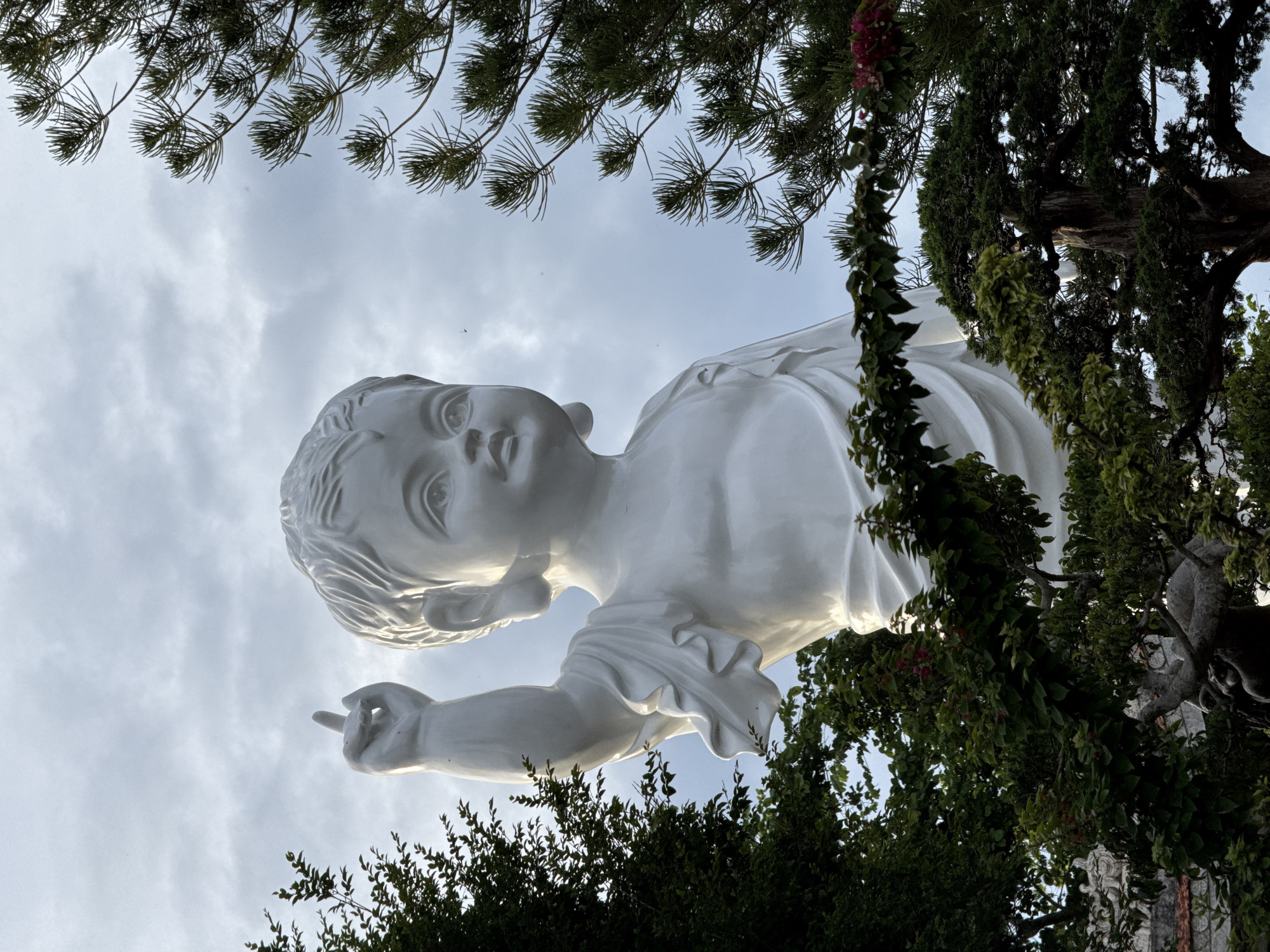 A low-angle shot depicts a white statue of a child, likely Jesus, pointing upward toward a cloudy sky. The statue is framed by tree branches and foliage. The photo was taken in Thôn 5, Vietnam.