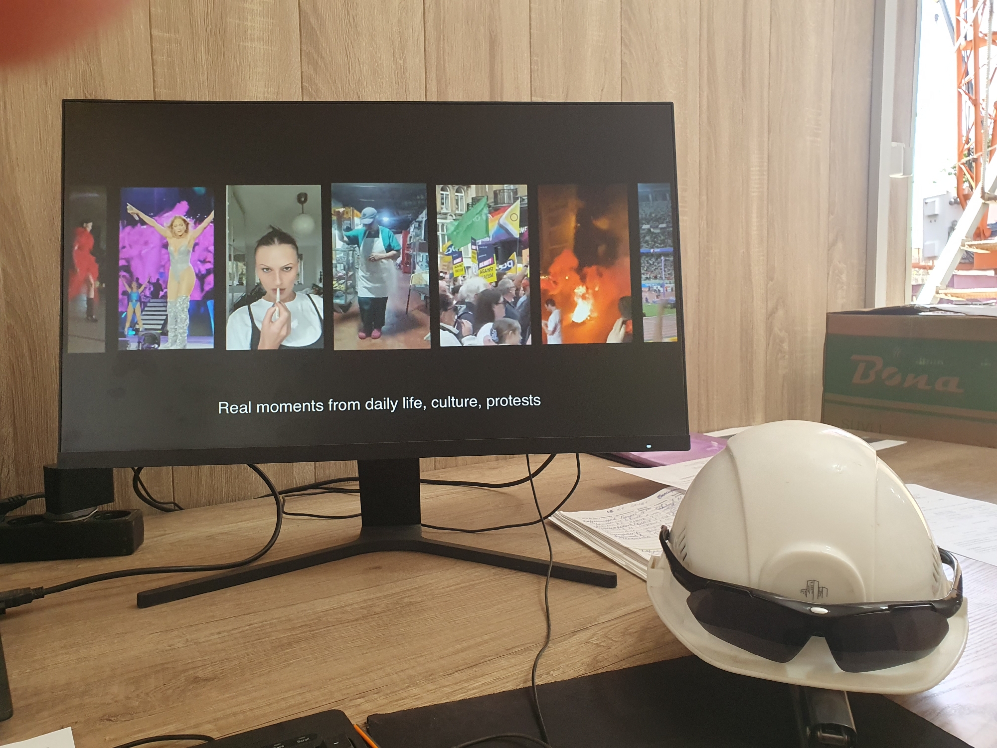 The image shows a desk with a computer monitor displaying a series of photographs. The photographs appear to be a collage of different scenes, including what looks like a concert or performance, a person holding a cigarette, a person in an apron, people at a protest with flags, a bonfire, and a sporting event. Below the photographs, text reads "Real moments from daily life, culture, protests".
In the foreground, on the desk, is a white hard hat with dark sunglasses resting on top. There are also papers, cables, and a keyboard visible on the desk. The background consists of a wooden paneled wall. A large window or opening is visible on the right side of the image, with scaffolding or construction equipment outside. A cardboard box with "Bona" written on it is placed near the window.
The scene does not contain any specific visual cues that would indicate its location in Tashkent, Uzbekistan.