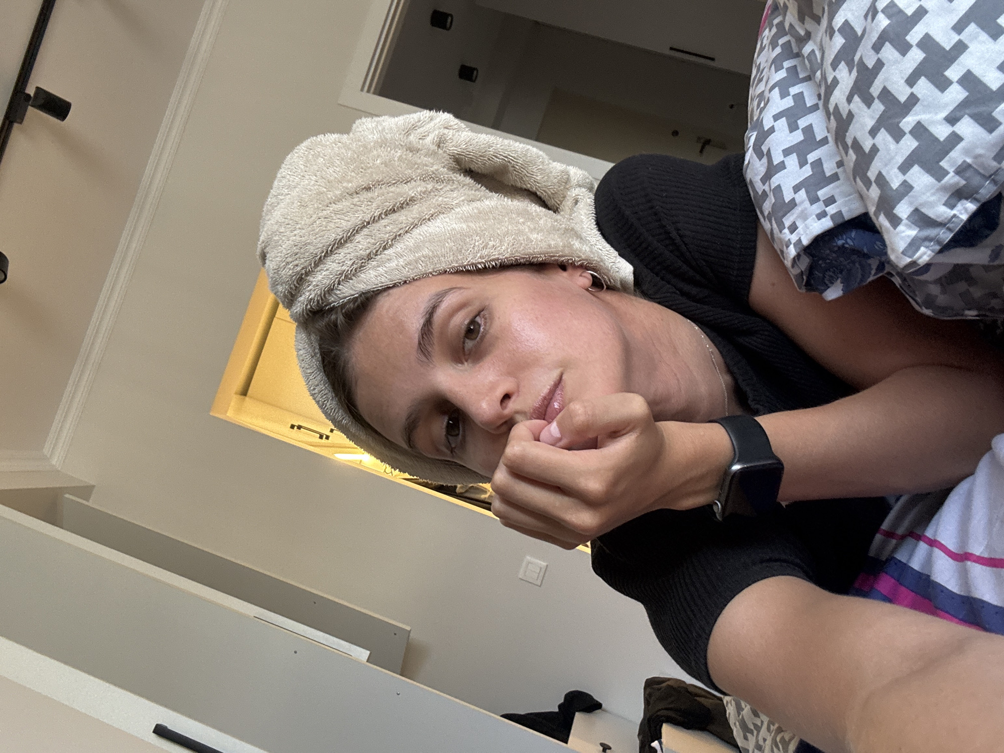 The image captures a person, likely female, lying down and looking directly at the camera, taking a selfie from a low angle. She has a light beige towel wrapped around her head, suggesting she may have recently washed her hair. She is wearing a black ribbed top or sweater and a black smartwatch on her left wrist. One hand is resting near her mouth, with her fingers lightly touching her lips, conveying a calm or thoughtful expression. Small hoop earrings are visible in her ears.
The background indicates an indoor setting, possibly a bedroom or living area. To the left, there are light-colored walls with white molding and black track lighting fixtures on the ceiling. In the upper background, a warm light emanates from what appears to be a window or a recessed light fixture, creating a soft glow. Light-colored shelves or furniture are visible in the lower left and center background, along with a white electrical outlet or switch on the wall.
Part of the bedding or a blanket with a distinct gray and white geometric pattern (resembling a houndstooth or similar motif) is visible to the right of the person's head. Below that, another piece of fabric with blue, pink, and white stripes can be seen. The overall impression is one of a relaxed, casual moment captured indoors. No visible text is present in the image.