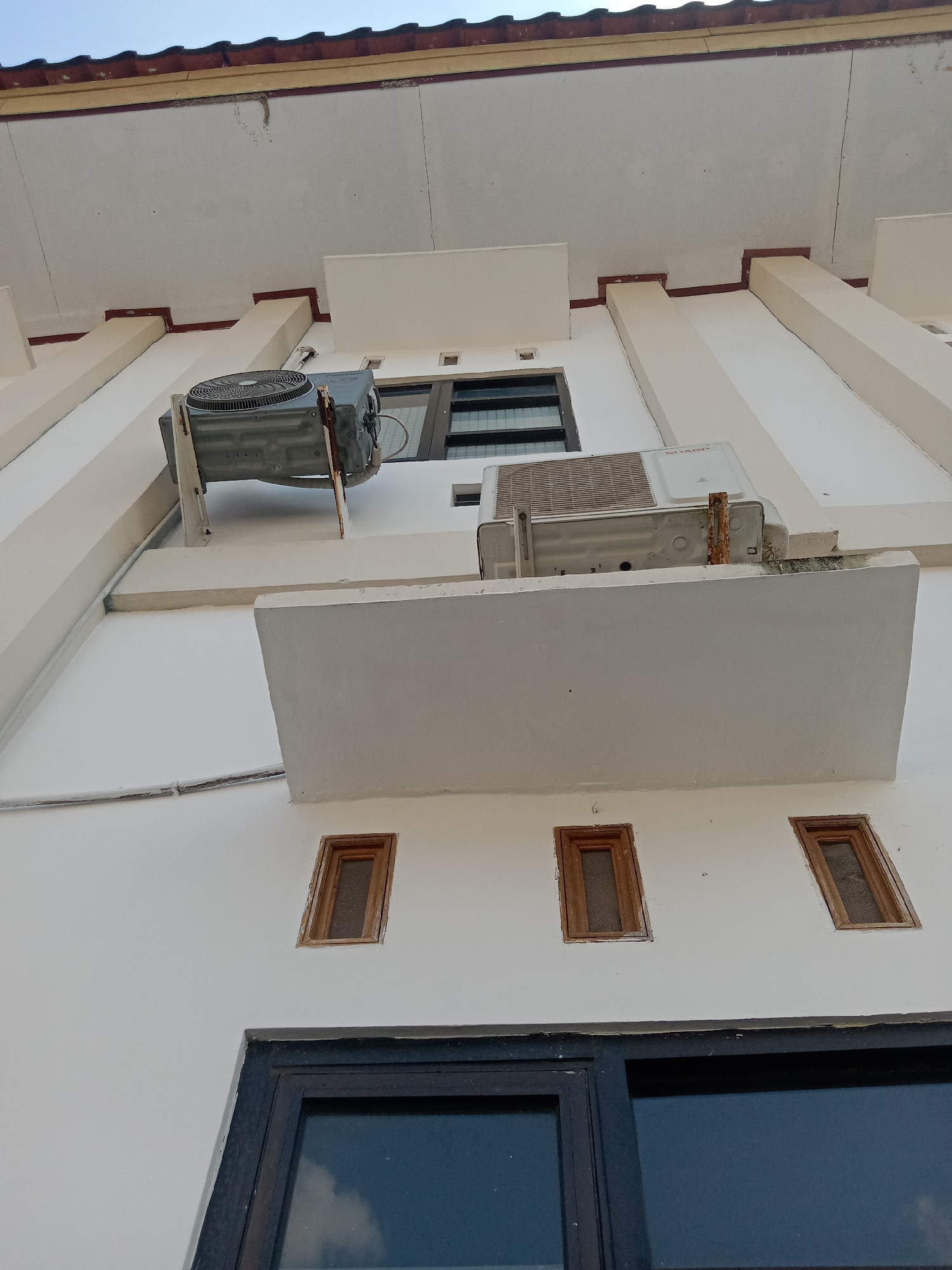 The image shows the exterior of a building, likely in the city of Klepek, Indonesia. The viewpoint is looking upwards from the ground, towards the building's facade. Two outdoor air conditioning units are visible, mounted on the wall at different heights. The upper unit is grey and appears to be older and slightly rusty. The lower unit is white and also shows signs of rust, particularly around its mounting bracket. Several small, rectangular windows with wooden frames are visible on the white wall below the air conditioning units. The building has a white facade, with some decorative architectural elements painted in a light beige color, and a dark tiled roofline at the top. A large dark-framed window or door is present at the bottom of the image. The overall impression is of a functional, multi-story building, possibly residential or commercial, with evidence of outdoor mechanical equipment.