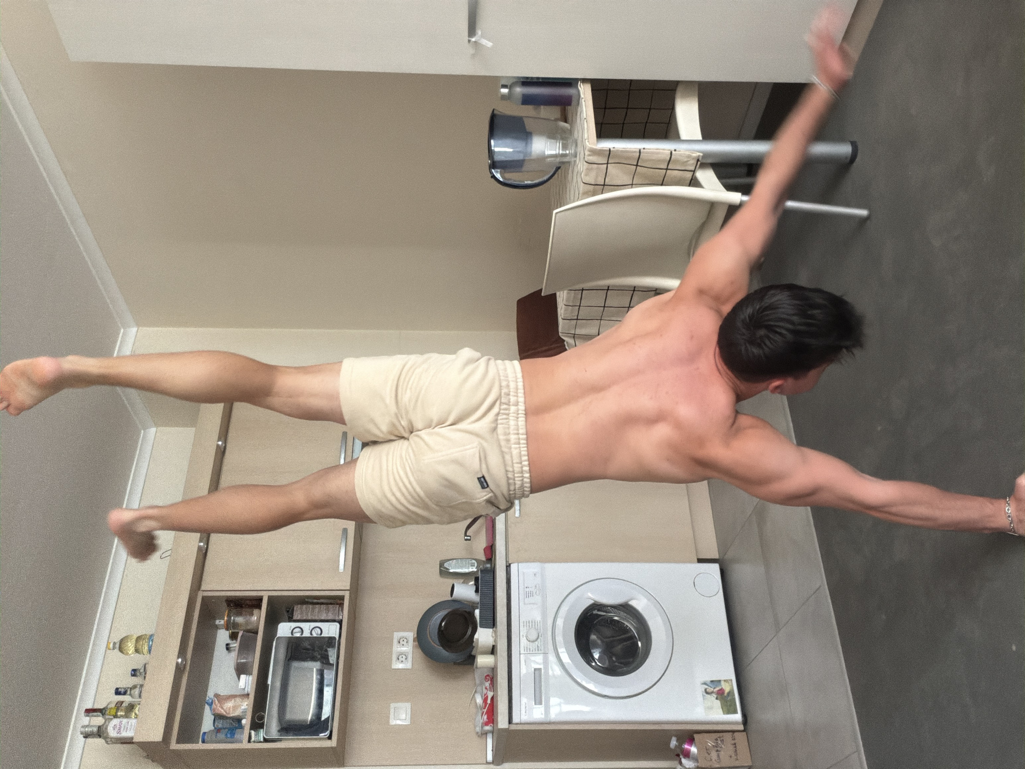 A man in beige shorts is performing a gymnastic pose. His right hand is pressed against a gray floor. His legs are extended to the left. The photo was taken in a room with beige walls. To the left, there is a kitchen unit with a microwave and various bottles. There is a white washing machine beneath the man. Behind him is a white chair and a shelf with a water filter jug. The location could be a modern apartment in Sofia, Bulgaria, given the architectural style and the types of appliances present.