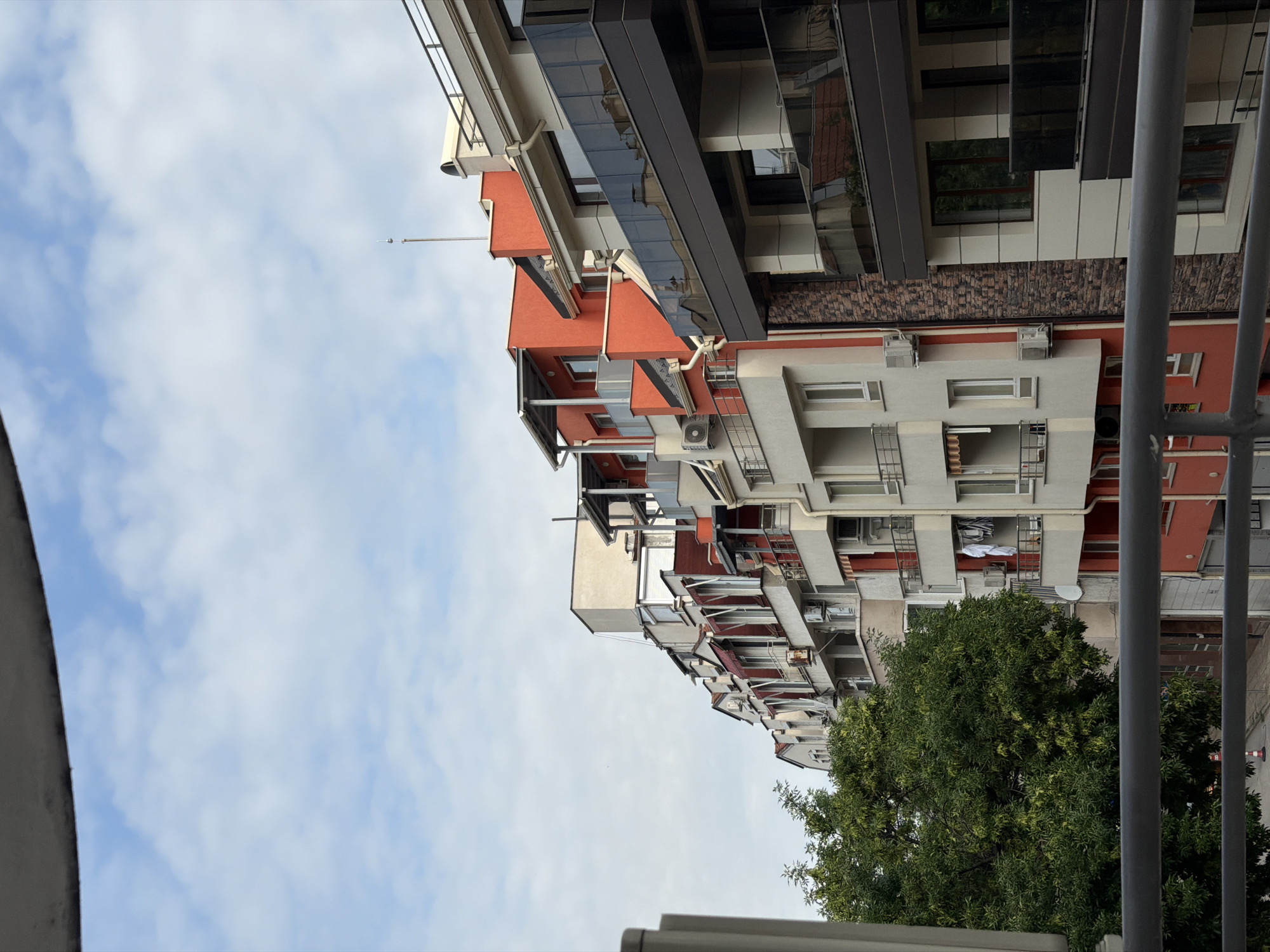 The image shows a row of apartment buildings in Plovdiv, Bulgaria, with a cloudy sky overhead. The buildings vary in architectural style and color, with some featuring red and white facades, while others have a more modern design with dark glass balconies. A large, leafy green tree is visible on the left side of the frame, and a metal railing runs along the bottom, suggesting the photo was taken from a balcony or similar elevated viewpoint. There are no people visible in the image. The scene depicts a residential urban environment.