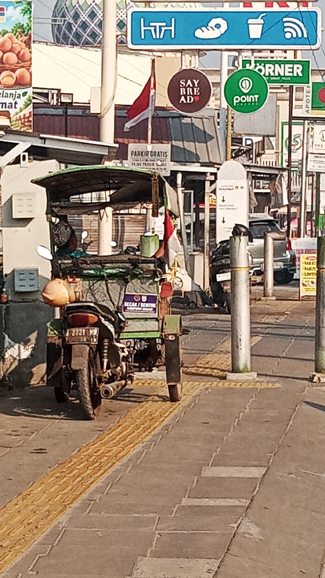 The image captures a street scene in Jawa, Indonesia, focusing on a motorized tricycle, known locally as a 'becak motor' or 'bentor.' The bentor is predominantly green with a canvas roof and appears to be transporting goods or passengers, though none are clearly visible. A large orange helmet rests on the side of the vehicle. The bentor is parked on a paved street with a textured yellow tactile paving strip running alongside the sidewalk.
In the background, several signs are visible. One prominent sign features the text "PARKIR GRATIS" (Free Parking) in bold letters. Above this, a red and white Indonesian flag is displayed. Other signs include a blue sign with icons representing food, drinks, and Wi-Fi, a circular brown sign with "SAY BRE AD," and a green circular sign with a white dot logo, likely indicating a business or service. A silver car is parked in the distance. The overall environment suggests a commercial or urban setting with a busy street and various establishments. The lighting indicates it is daytime, with sunlight casting shadows on the ground.