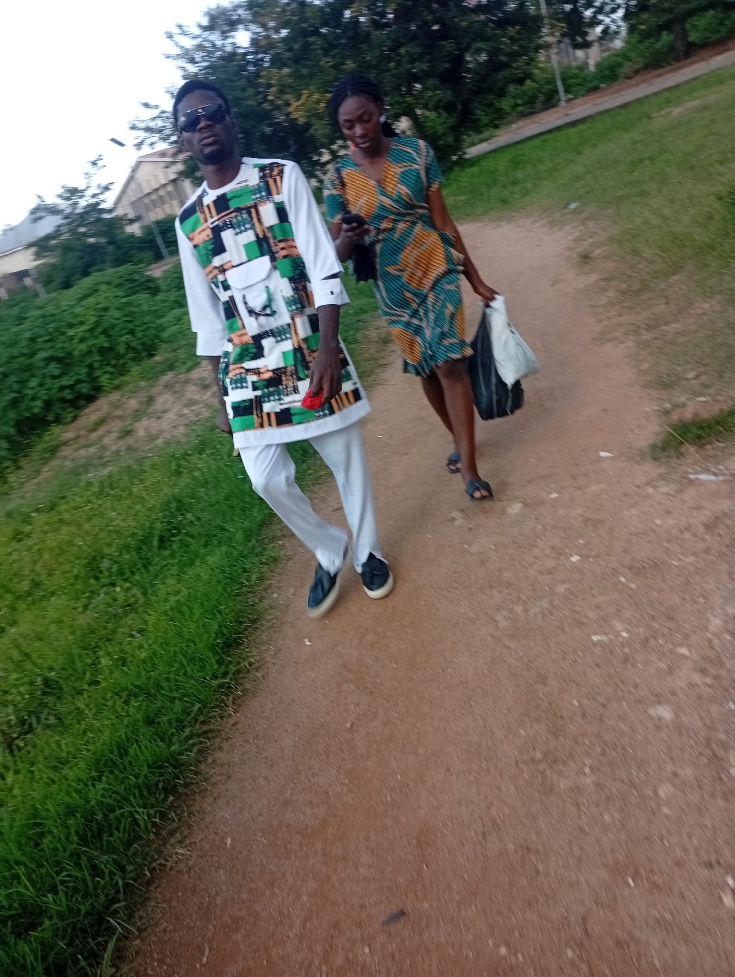 Two individuals, a man and a woman, are walking on a dirt path in what appears to be a park or a less developed area of the city of Jos, Nigeria. The man, in the foreground, is wearing a white outfit with a colorful, geometric-patterned tunic and white pants. He has on dark sunglasses and is holding a red object in his right hand. The woman, walking behind him, is wearing a short, patterned dress in shades of green and orange, and is carrying two shopping bags, one white and one black, in her left hand. She is looking down at her phone. The path is bordered by lush green grass on the left and trees and foliage in the background. A building, possibly a residential or administrative structure, is visible through the trees on the left side of the image. The overall lighting suggests it is either early morning or late afternoon.
