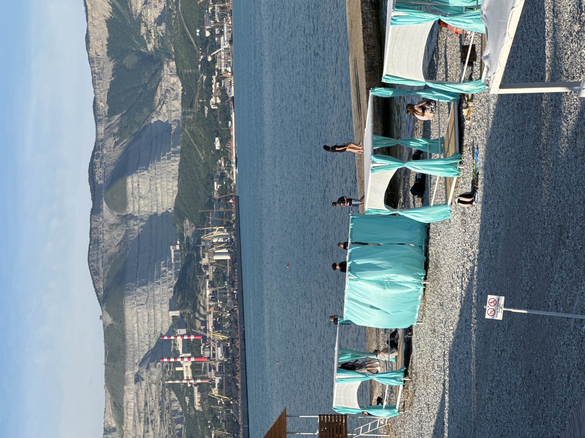 The image depicts a view of the beach and sea in Novorossiysk, Russia, with a backdrop of industrial facilities and a large, terraced quarry on the mountainside. In the foreground, a pebble beach is lined with several white and turquoise shaded beach cabanas. Some people are visible relaxing in or around these cabanas. A sign prohibiting cycling is planted in the beach. The sea stretches across the midground, with a concrete pier extending into it. Several boats are visible on the water, and in the distance, industrial structures including cranes and smokestacks are present near the shoreline, with the prominent, layered slopes of the quarry dominating the upper portion of the scene. The sky is clear and blue.