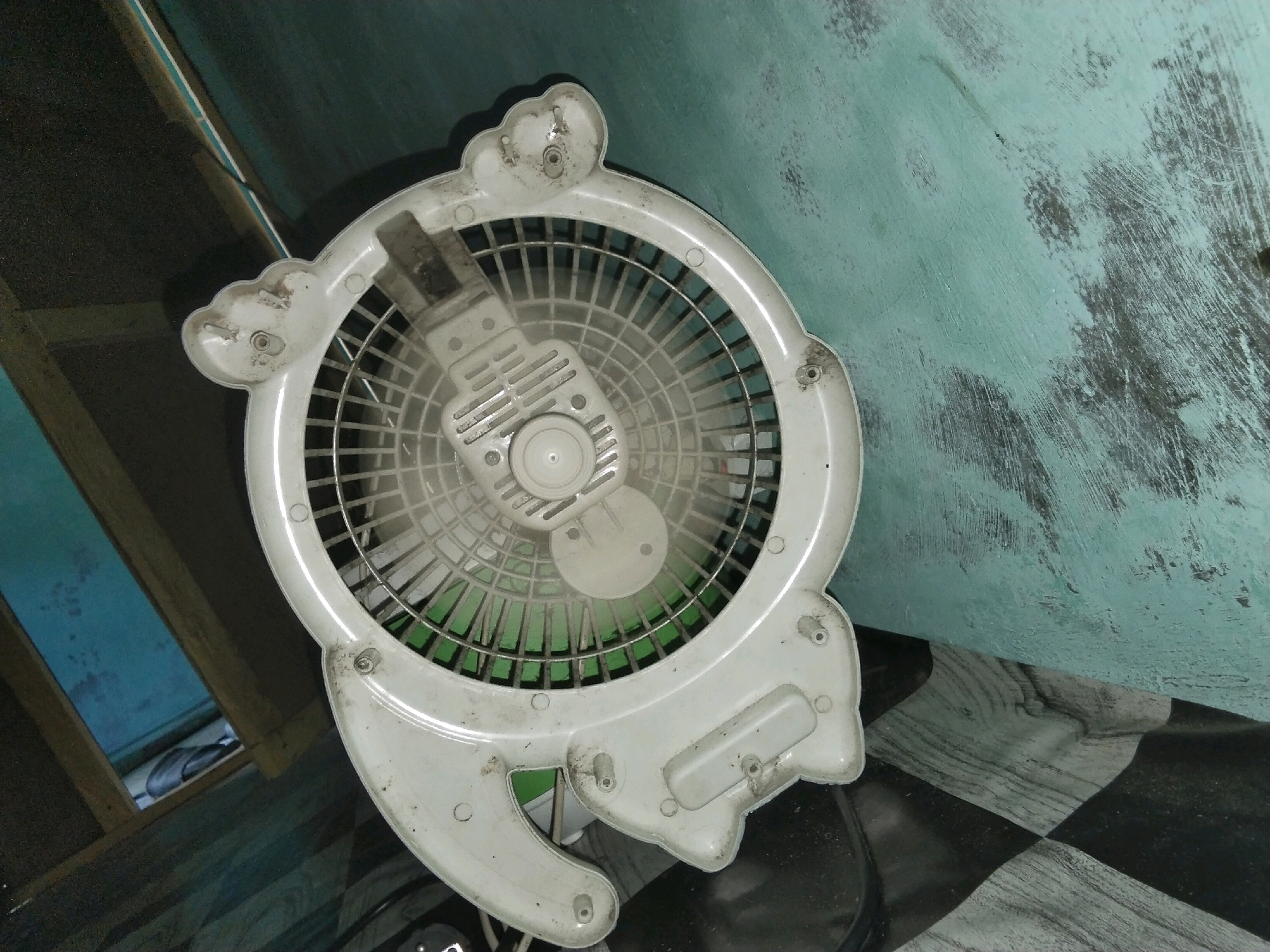 In Moro, Indonesia, a white electric fan with a cage is pictured resting on a checkered floor. The fan exhibits a cartoonish design, resembling a cat's face with two rounded protrusions at the top and two paw-like structures at the bottom. The fan's casing is dusty, with visible grime and some dirt accumulation around the screws and edges. The fan's blades are a light green color. The background features a distressed turquoise wall with visible patches of darker undercoat showing through, suggesting wear and tear. To the left of the fan, a wooden structure is visible, partially obscuring a glimpse of what appears to be a pair of flip-flops. The lighting in the scene is somewhat dim.