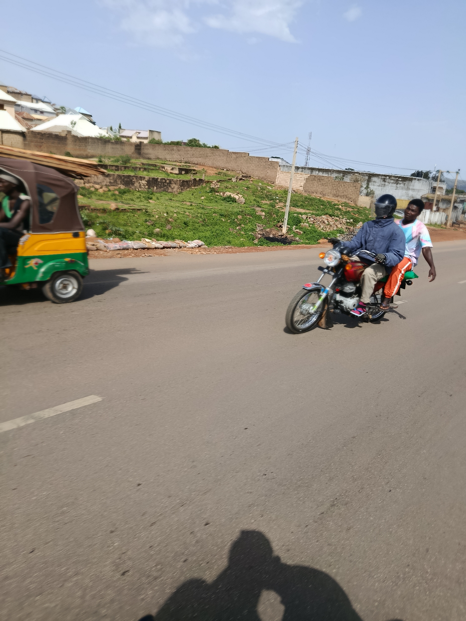 An eye-level, medium shot captures a street scene in Jos, Nigeria, under a bright, partly cloudy sky. On the right, two individuals are riding a red motorcycle. The driver, wearing a dark blue jacket and a black helmet, is positioned in front. Behind him, a passenger in a multi-colored shirt and orange pants leans forward. The motorcycle is in motion, indicated by the blurred background and the slight tilt of the vehicle.
To the left of the motorcycle, a yellow and green auto-rickshaw, also in motion, is visible. Its canvas roof is brown. A person in a green shirt is seated inside the rickshaw. The vehicle appears to be traveling in the same direction as the motorcycle.
The road is paved with asphalt, and a white line marks the lane. To the left and behind the vehicles, a gently sloping hill is covered in lush green grass and scattered rocks. A brick wall runs along the mid-section of the hill. Above the wall, several buildings with various roof colors, including white, brown, and blue, are visible. Power lines crisscross the upper part of the image. The overall scene suggests a typical day on the roads of Jos, characterized by common modes of transportation and the city's hilly terrain.