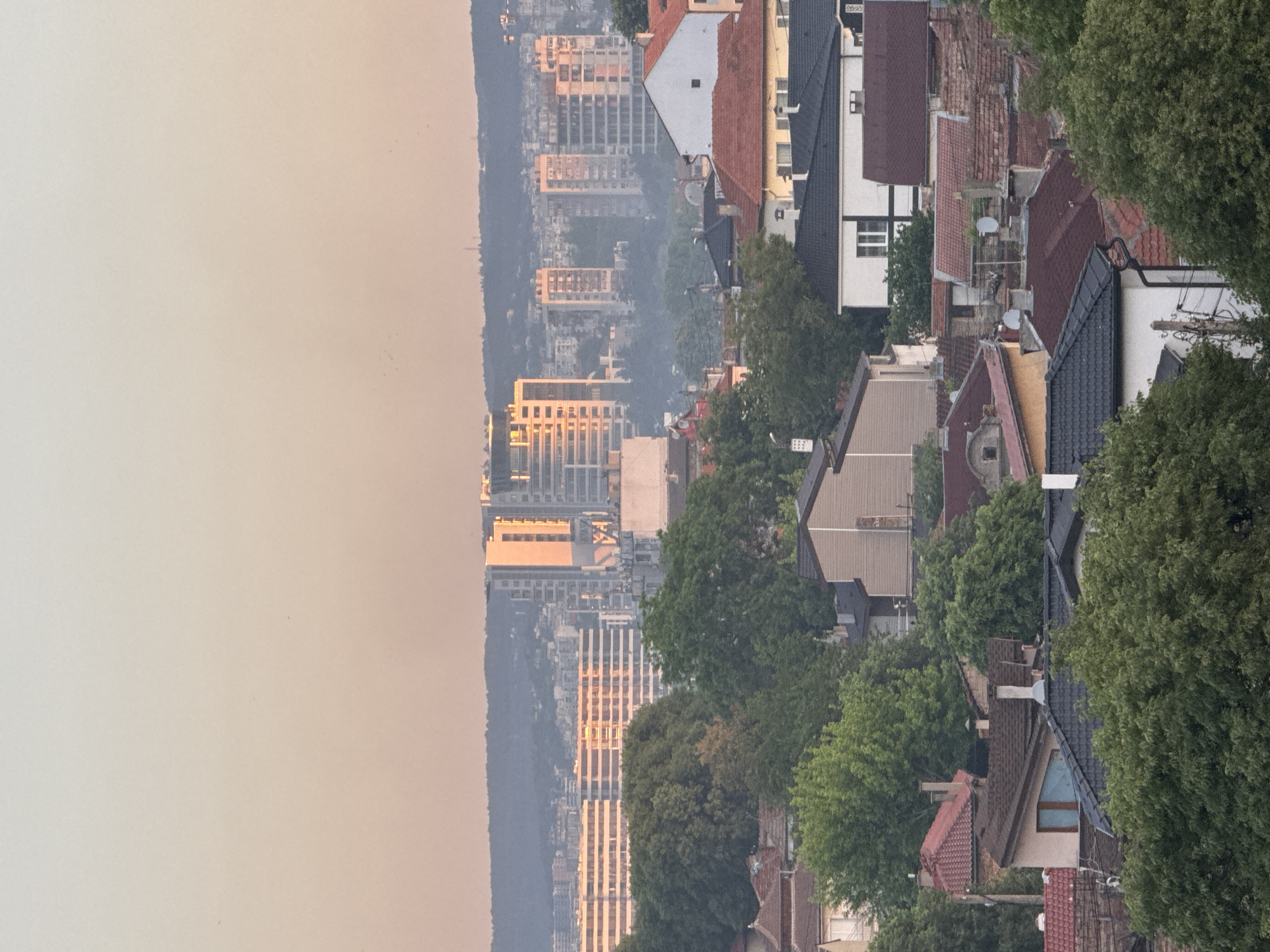A high-angle view shows a cityscape in Varna, Bulgaria, juxtaposing residential rooftops with distant high-rise buildings. The foreground is characterized by a mix of tiled roofs in various shades of red, brown, and grey, interspersed with lush green trees. Some houses have visible windows and chimneys. In the background, tall, modern buildings reflect the sunlight, partially obscured by what appears to be smog or a dusty haze. The sky, primarily visible on the left, is a pale orange, likely indicating either sunrise or sunset. The overall scene depicts a blend of urban and natural elements.