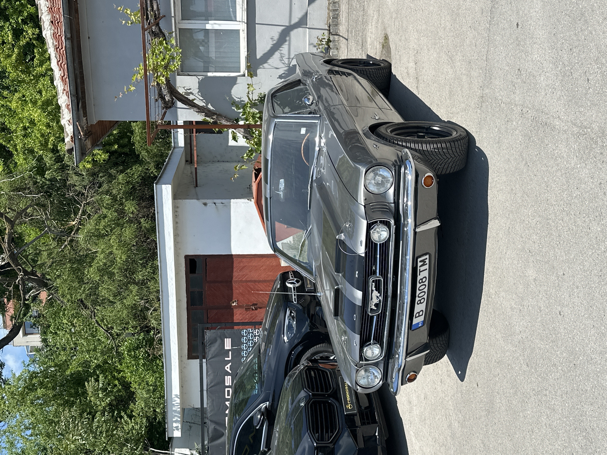 The image shows a gray classic Ford Mustang parked on a concrete surface in what appears to be an urban environment. A black BMW SUV is partially visible in front of the Mustang, partially blocking the view of the Mustang. Behind the cars is a building with a partially visible red door and a black sign that appears to say "MOSALE". Green foliage is also present near the building. The Mustang has a license plate with the code "B 8008 TM", indicating it is likely in Bulgaria. A window on the building shows an outdoor terrace area with plants growing. The setting appears to be a sunny day. It's impossible to verify the exact location in Varna, Bulgaria.