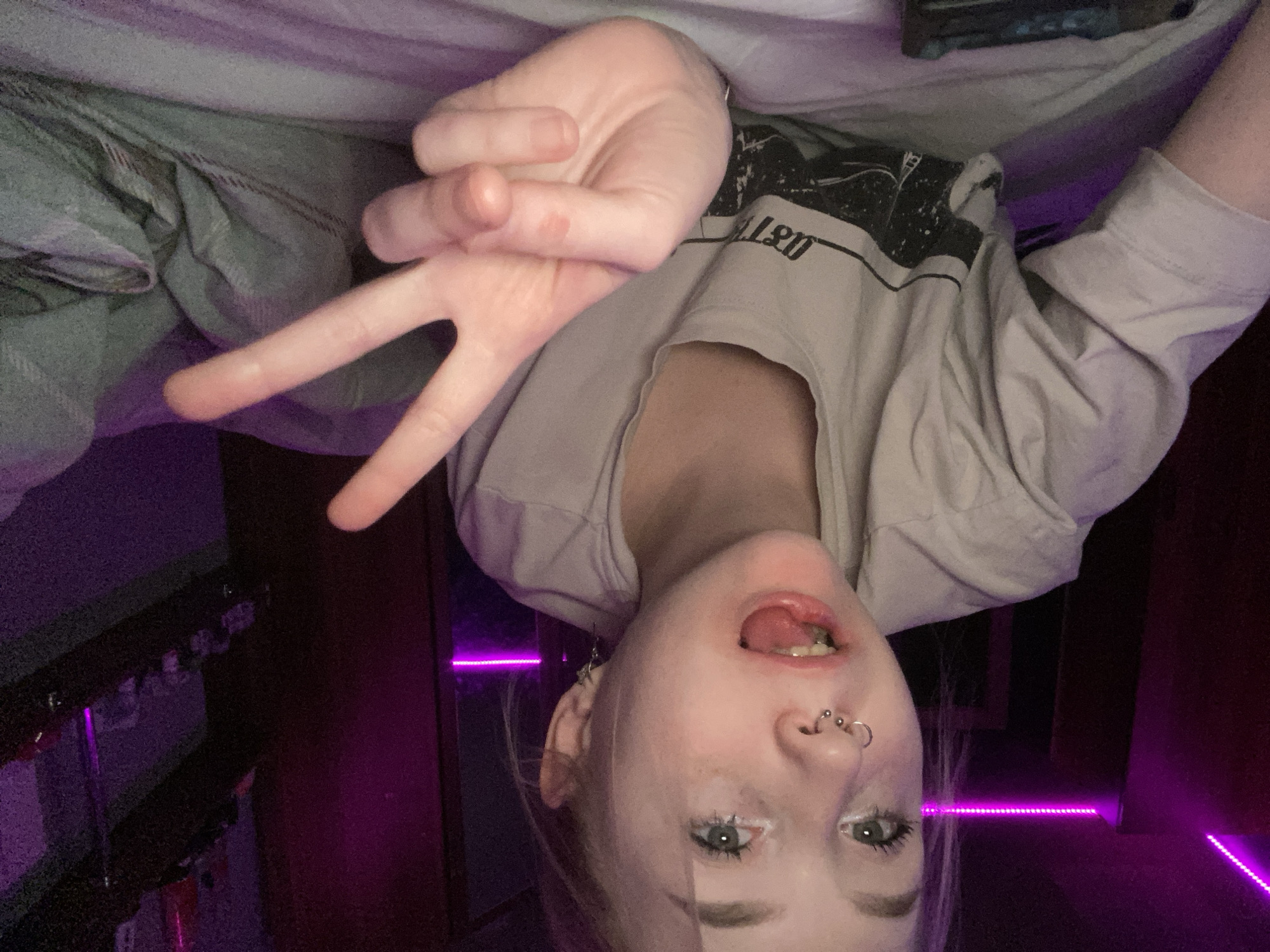 The image depicts a young individual, likely female, with light hair and eyes, a nostril ring, and a septum piercing, lying face down on a bed. The person is wearing a light-colored, short-sleeved t-shirt featuring a dark graphic design on the chest. Their mouth is open with the tongue slightly extended, and they are looking directly at the camera. Their right hand is raised, performing a two-finger "peace" sign gesture. The background reveals a dimly lit room, illuminated primarily by a horizontal strip of vibrant purple LED lighting across the upper wall. To the right, a dark shelving unit is visible, holding indistinct items. The bedding under the individual appears to be a mix of light grey and white. A dark, rectangular object, possibly an electronic device, rests on the bedding in the lower left foreground. The specific location within Izhevsk, Russia, cannot be determined from the visual information provided.
