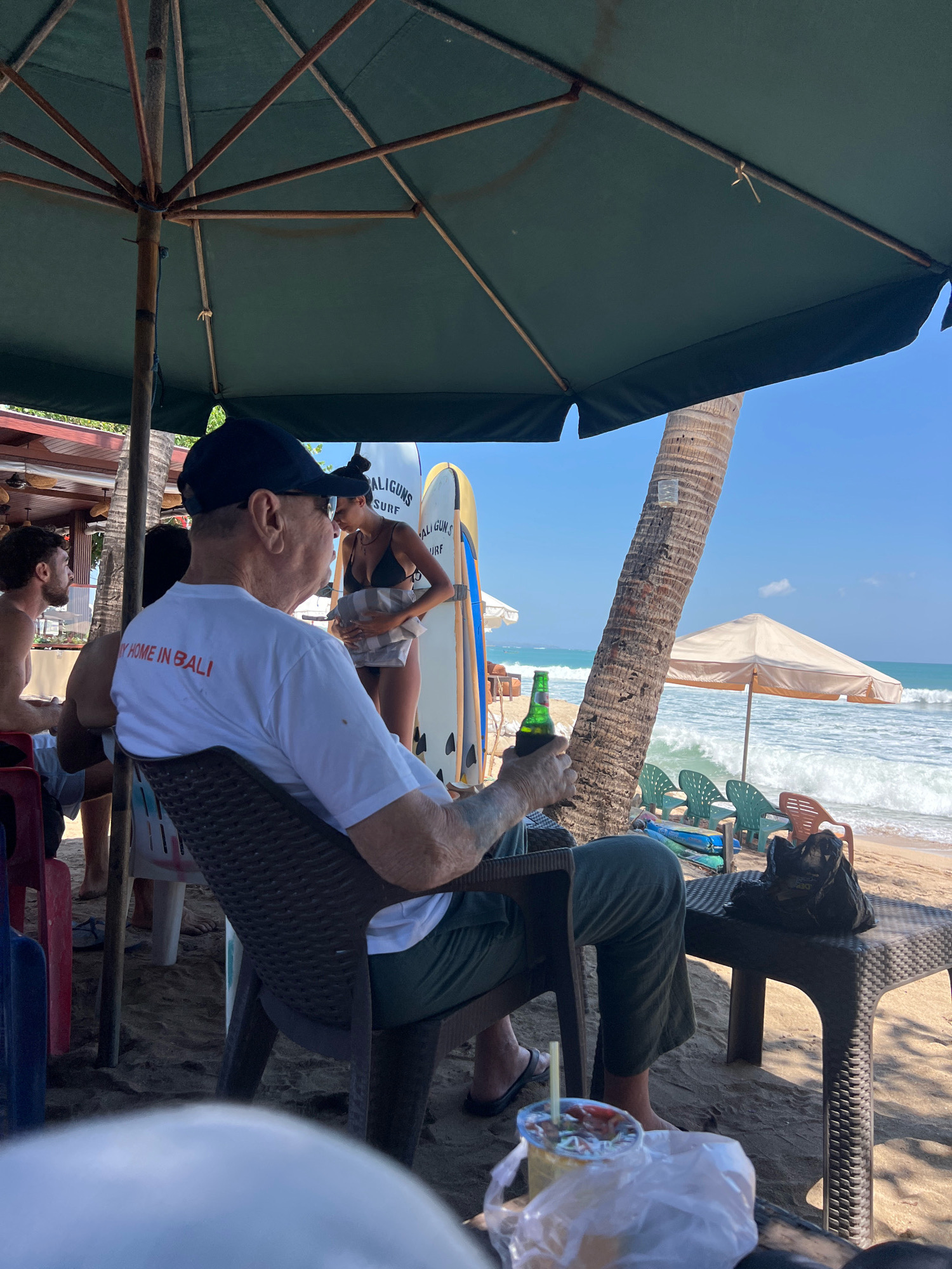 The image depicts a beach scene in Kuta, Indonesia. In the foreground, an elderly man wearing a white t-shirt with red text that reads "MY HOME IN BALI" and a dark blue baseball cap is seated in a brown plastic chair. He is holding a green glass bottle of beer in his right hand. In front of him, on a dark plastic table, is a drink with a straw and a white plastic bag.
Behind the man, a woman in a black bikini stands next to two surfboards. She is holding a white towel. To the left, partially visible, is a man with dark hair and a beard. The background shows the beach with waves, several beach chairs, and a beach umbrella. A palm tree stands to the right of the man. The overall lighting suggests it is a sunny day.