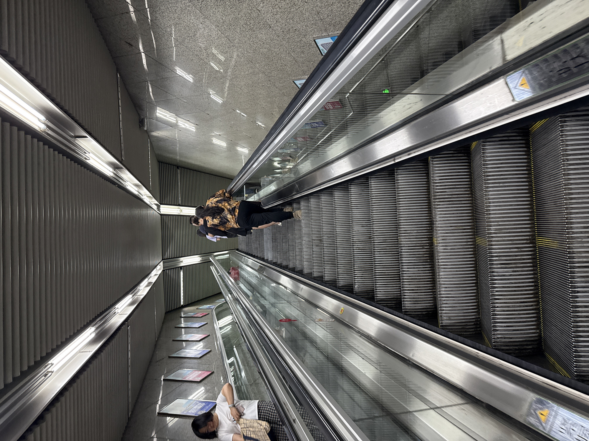 The image, taken in 龙溪街道, China, shows a perspective view from above of a set of escalators within what appears to be a subway or underground transportation hub. Two escalators are visible. One is operating and has two people riding it, while the other has one person lying on it. The surroundings include architectural details such as walls with linear patterns, lighting fixtures, and tiled flooring. There are posters or advertisements fixed on the walls.