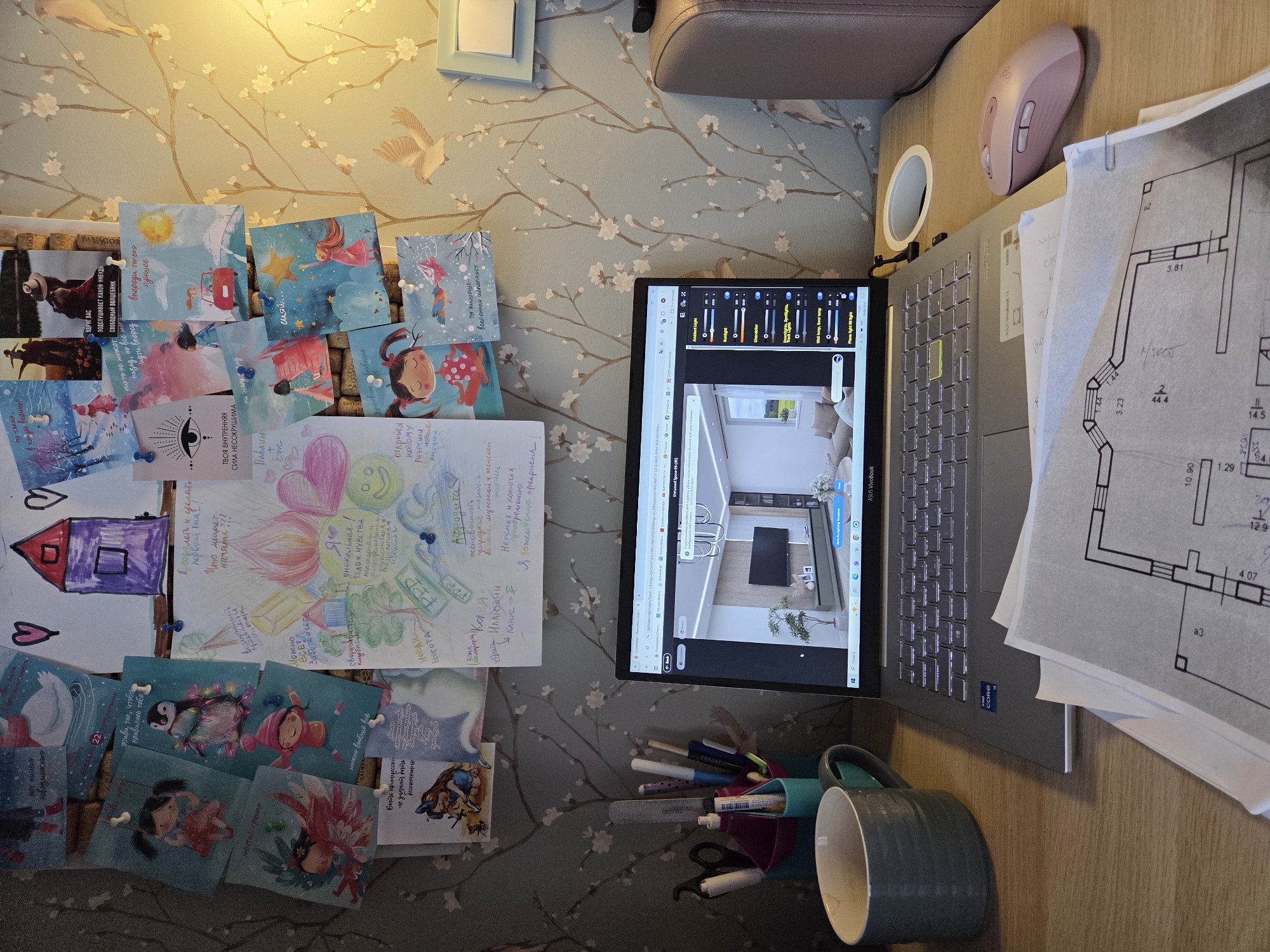 The image shows a cluttered desk with a laptop, blueprints, and various decorative items. The laptop displays a 3D interior design rendering of a living room, with lighting controls visible on the right side of the screen. A pink wireless mouse is positioned to the right of the laptop. To the left of the laptop, a grey mug and a holder filled with pens and markers are visible. Above the desk, a wall is decorated with numerous pinned photos, drawings, and inspirational quotes. The overall setting appears to be a home office or study area. The image does not provide any specific information about the location in Birobidzhan, Russia.