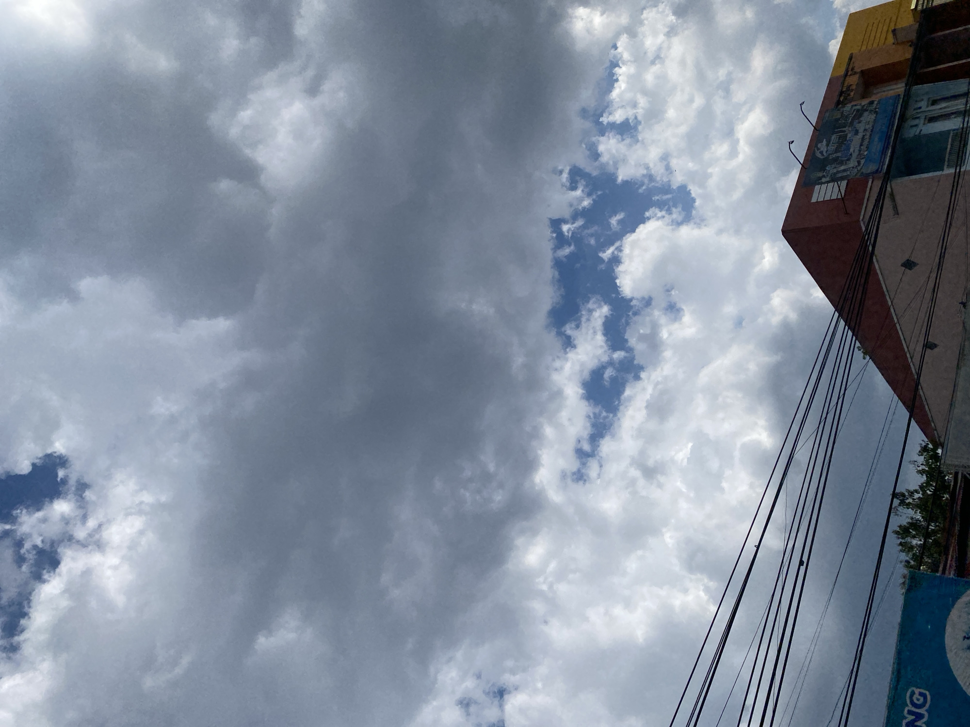 The image is a low-angle, upward shot of a sky filled with a mixture of dark and light gray clouds, with patches of blue sky visible. In the foreground, several black electrical wires are strung across the frame. Below the wires, the upper parts of buildings are visible. One building on the right has a prominent red, angular roof and a colorful facade featuring a large mural. To the left, a portion of a blue sign with white text is visible, and trees can be seen behind the buildings. The scene suggests an urban environment in Banjarbaru, Indonesia, with a cloudy sky and overhead infrastructure.