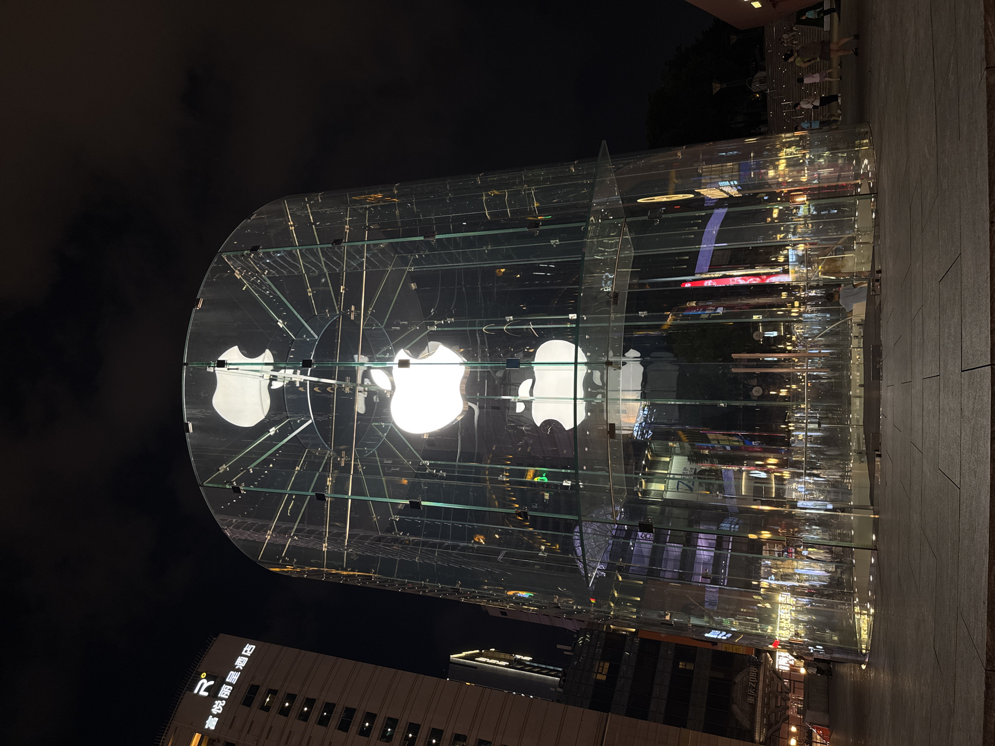 The image depicts an Apple Store with a distinctive cylindrical glass exterior, illuminated at night. Three large, white Apple logos are visible vertically stacked within the structure. The store is situated in a modern urban environment, likely a commercial district, as evidenced by the adjacent buildings, including a tall hotel with a prominent logo and Chinese characters that read "富悦丽景酒店" (Fuyue Lijing Hotel). The ground level around the store is paved and appears to be a public plaza. Several individuals are present in the scene, with some walking or standing near the store and others in the background, suggesting general pedestrian activity. The overall scene is characterized by artificial lighting, with the store itself being the most brightly lit element against the dark night sky. The architecture and the presence of the Apple Store indicate a contemporary urban setting in China. The specific location, as identified, is the Yuzhong District in China, likely within a commercial or shopping area.