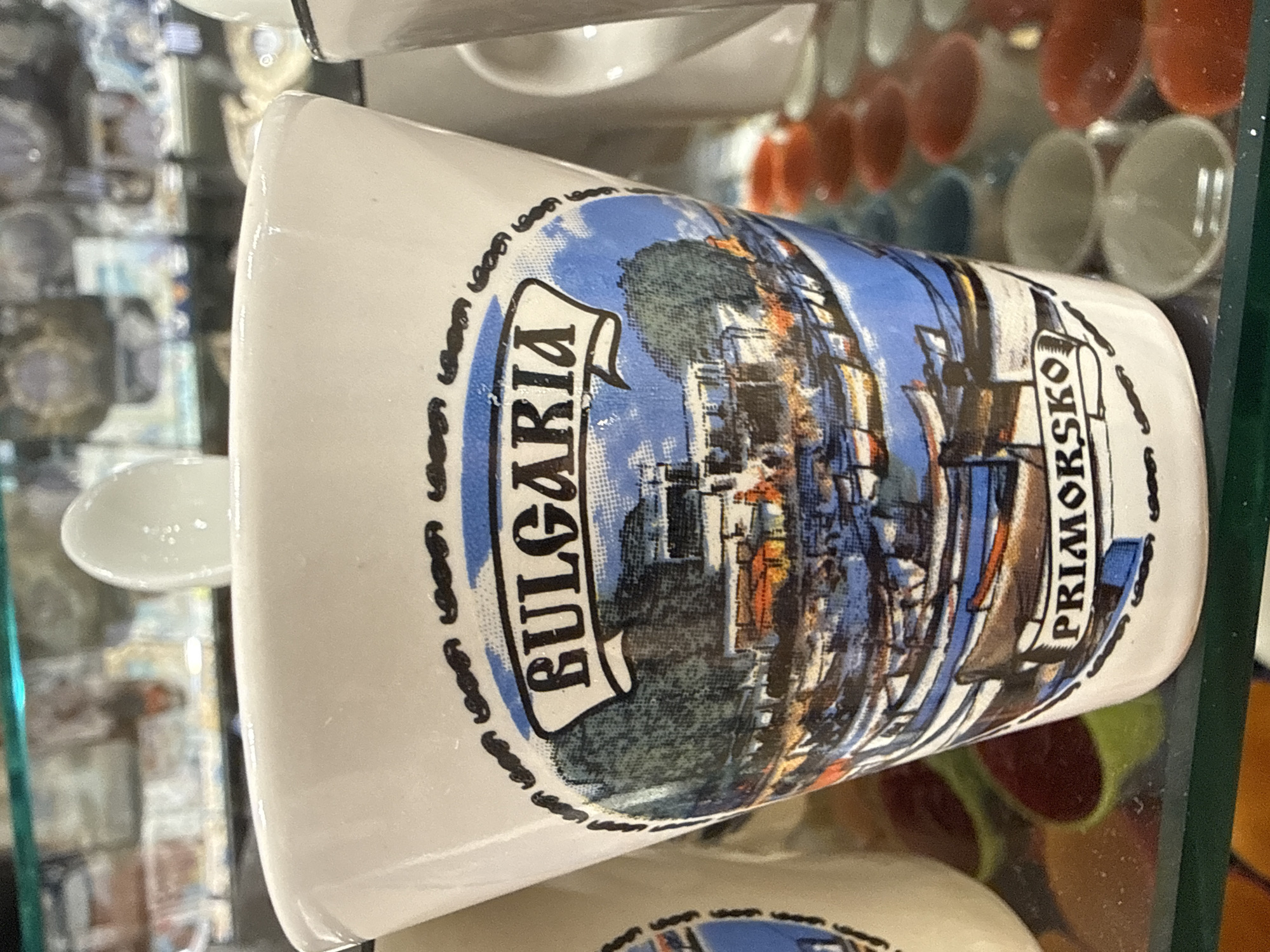 A white ceramic drinking vessel, possibly a shot glass or small mug, with a tapered body and slightly flared rim, is positioned on a transparent glass shelf. A small, white spoon-like object is visible directly behind the rim. The vessel features a multi-colored illustration framed by a black, ornate border with repetitive wavy motifs. The upper banner within the frame displays the word "BULGARIA" in prominent, black serif capital letters. The central illustration depicts a vibrant coastal scene: a body of blue water in the foreground and midground contains numerous small, colorful fishing boats. Along the distant shore, a cluster of buildings with visible red/orange roofs and light-colored walls is nestled against a backdrop of green trees or hills, under a blue sky with faint white clouds. The lower banner within the frame presents the word "PRIMORSKO" in black serif capital letters. The text explicitly identifies the location as Primorsko, a town in Bulgaria, and the image depicts its harbor or coastal area. To the right of the main vessel, another white ceramic mug with a visible handle is partially obscured. The glass shelf below the vessel displays subtle dust or speckles. Beneath the shelf, various colorful, amorphous shapes in red, green, and brown are visible. The background, largely out of focus, reveals indistinct items through what appears to be a glass display case, suggesting a retail or exhibition setting.