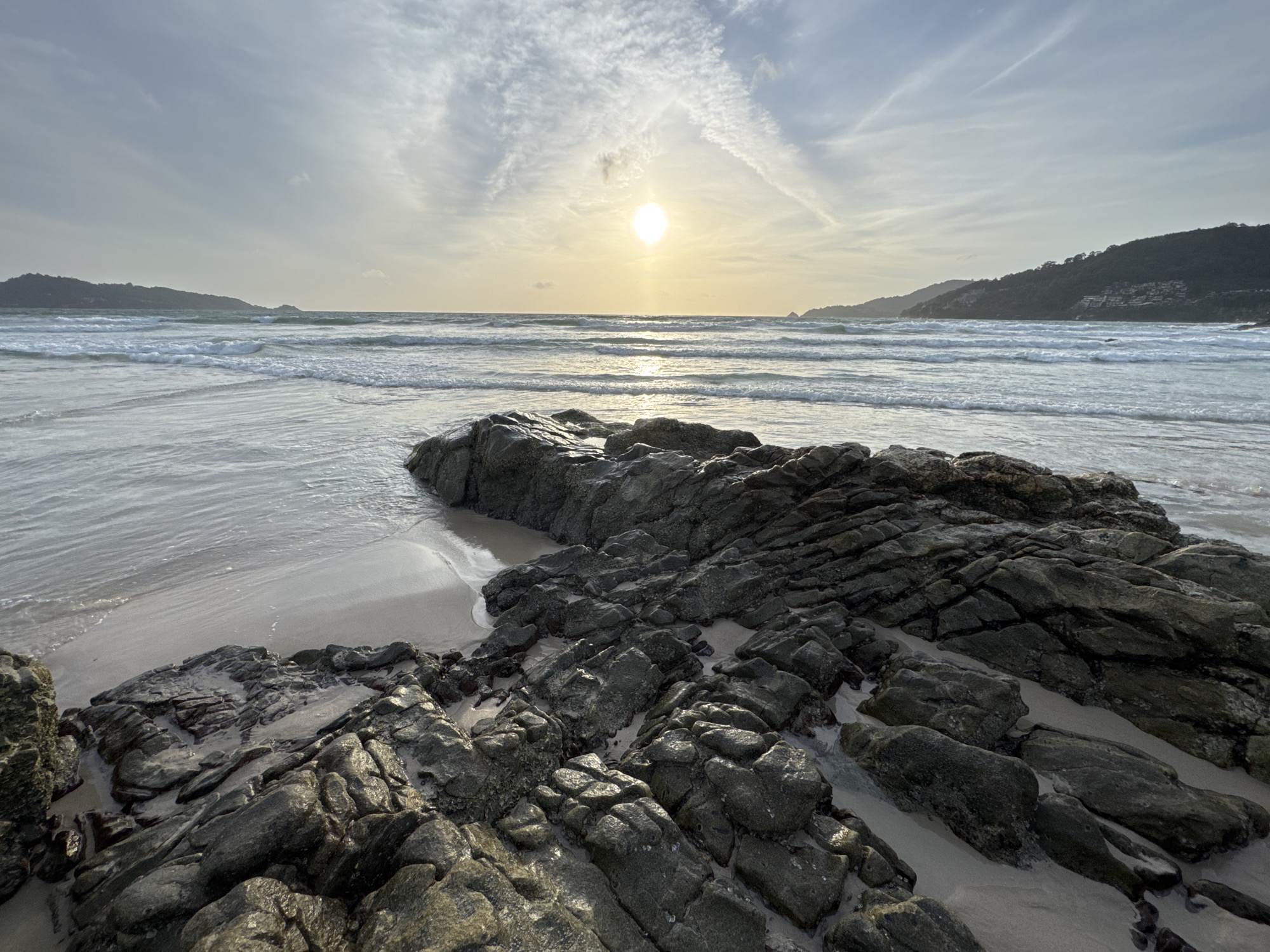 The image depicts a coastal scene at Patong Beach, Thailand, during either sunrise or sunset, as indicated by the low-hanging sun casting a warm glow. The foreground is dominated by dark, wet rocks and boulders scattered on the sandy shore. The tide is low, revealing the textures and crevices of the rocks, with some shallow pools of water reflecting the sky. Beyond the rocks, the gentle waves of the Andaman Sea lap onto the wet sand. In the middle ground, the sea stretches out with a series of white-capped waves rolling towards the shore. The horizon is marked by the silhouette of distant hills or islands on either side of the frame, covered in lush vegetation. The sky is a blend of soft blues and grays, with wispy clouds illuminated by the sun. The overall atmosphere is tranquil and serene, characteristic of a tropical beach at dawn or dusk. There are no people visible in the image.