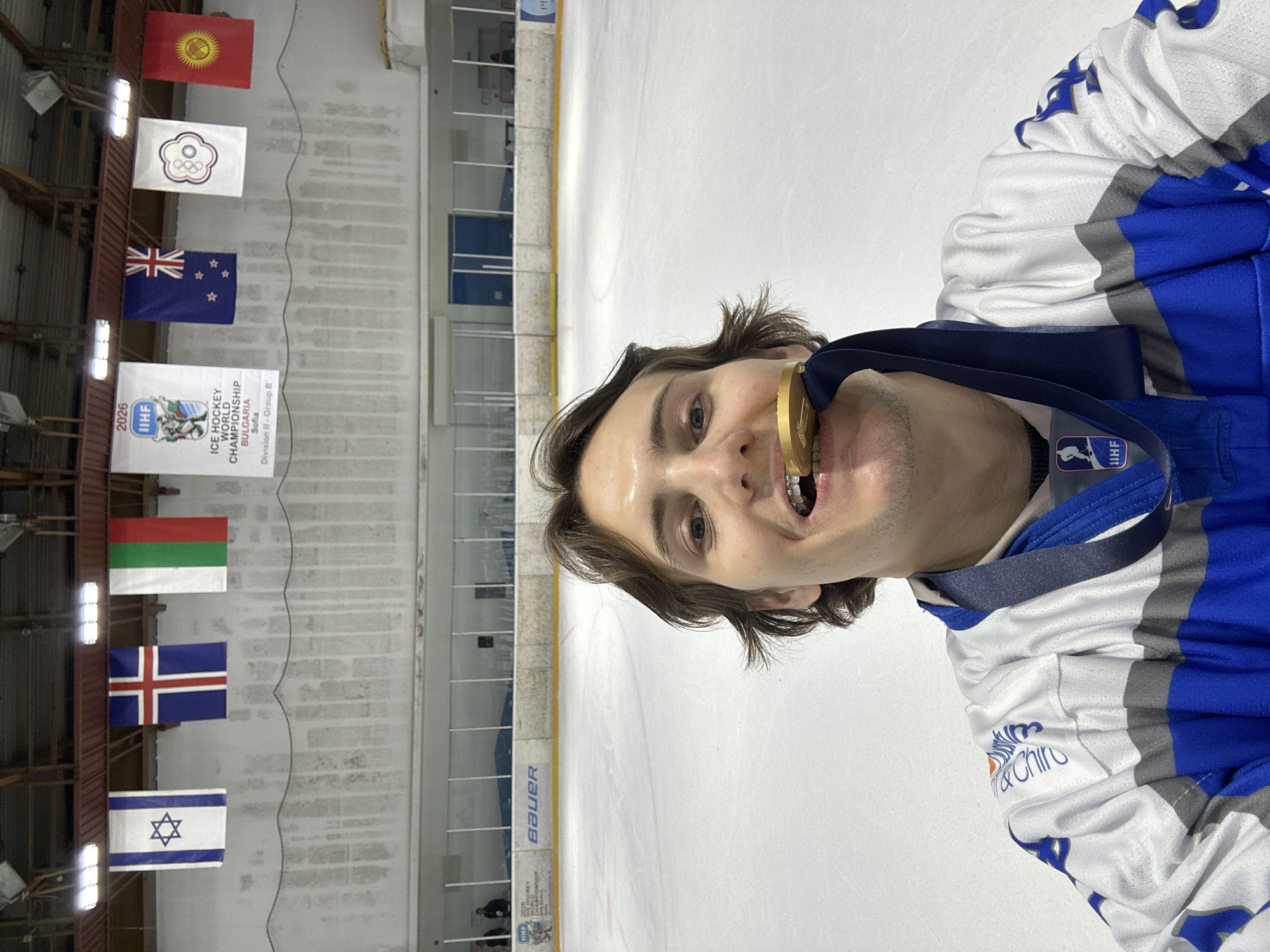 A man is on an ice rink. He is wearing a hockey jersey with a logo on the chest, and he has a gold medal in his mouth. Behind him are several flags and a banner that reads "2026 IIHF Ice Hockey World Championship, Bulgaria, Sofia, Division II - Group B". The flags include Kyrgyzstan, Chinese Taipei, New Zealand, Bulgaria, Iceland and Israel.