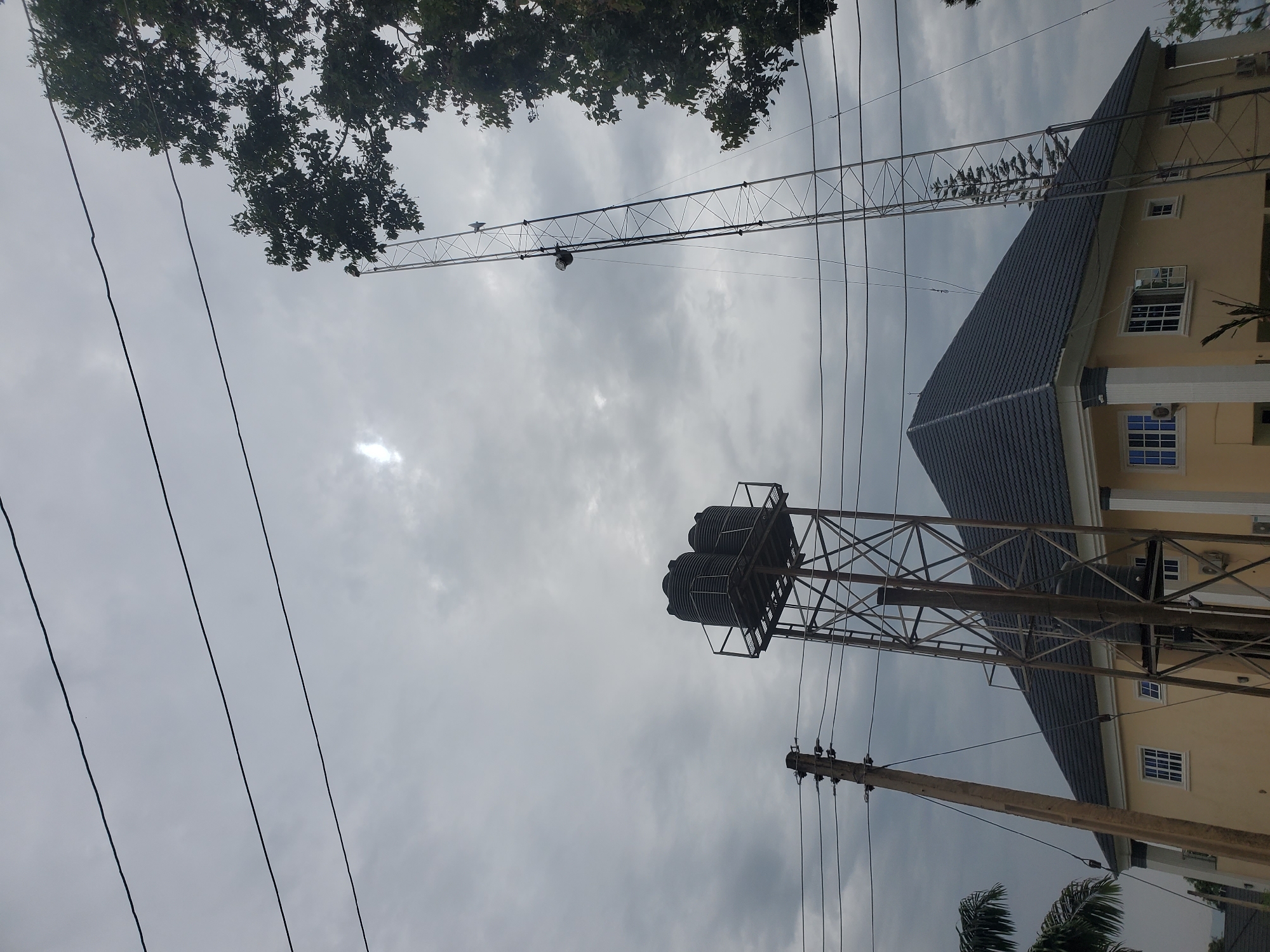 The image depicts an overcast sky over a residential building in Jos, Nigeria. A two-story cream-colored building with a dark gray tiled roof is visible, featuring white-trimmed windows. To the left of the building, a tall metal tower supports two large black water tanks. To the right, another taller, slender metal tower, possibly a communication mast, extends upwards. Electrical wires are strung across the frame, supported by wooden poles. A large tree with dark green foliage is situated on the far right, partially obscuring the taller tower. The overall scene is one of a cloudy day in a semi-urban or suburban environment in Jos.