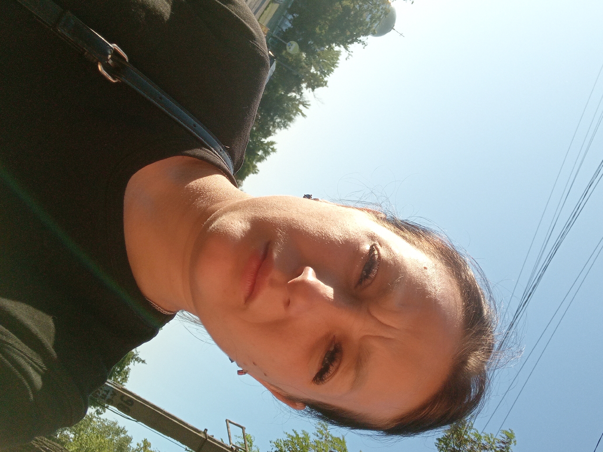 A selfie is taken by a woman with her face contorted in an expression of mild disgust or annoyance. The camera is angled upwards, capturing her forehead furrowed and her eyes looking sideways. She is wearing a black t-shirt and a dark strap of a bag crosses her chest. In the background, a clear blue sky dominates, with the tops of green trees visible on the right side of the frame. Some power lines are also visible in the upper left quadrant. The lighting suggests it is daytime, with sunlight casting shadows on her face. A dome structure with a spire is partially visible at the bottom of the image, hinting at a location with some architectural landmarks.