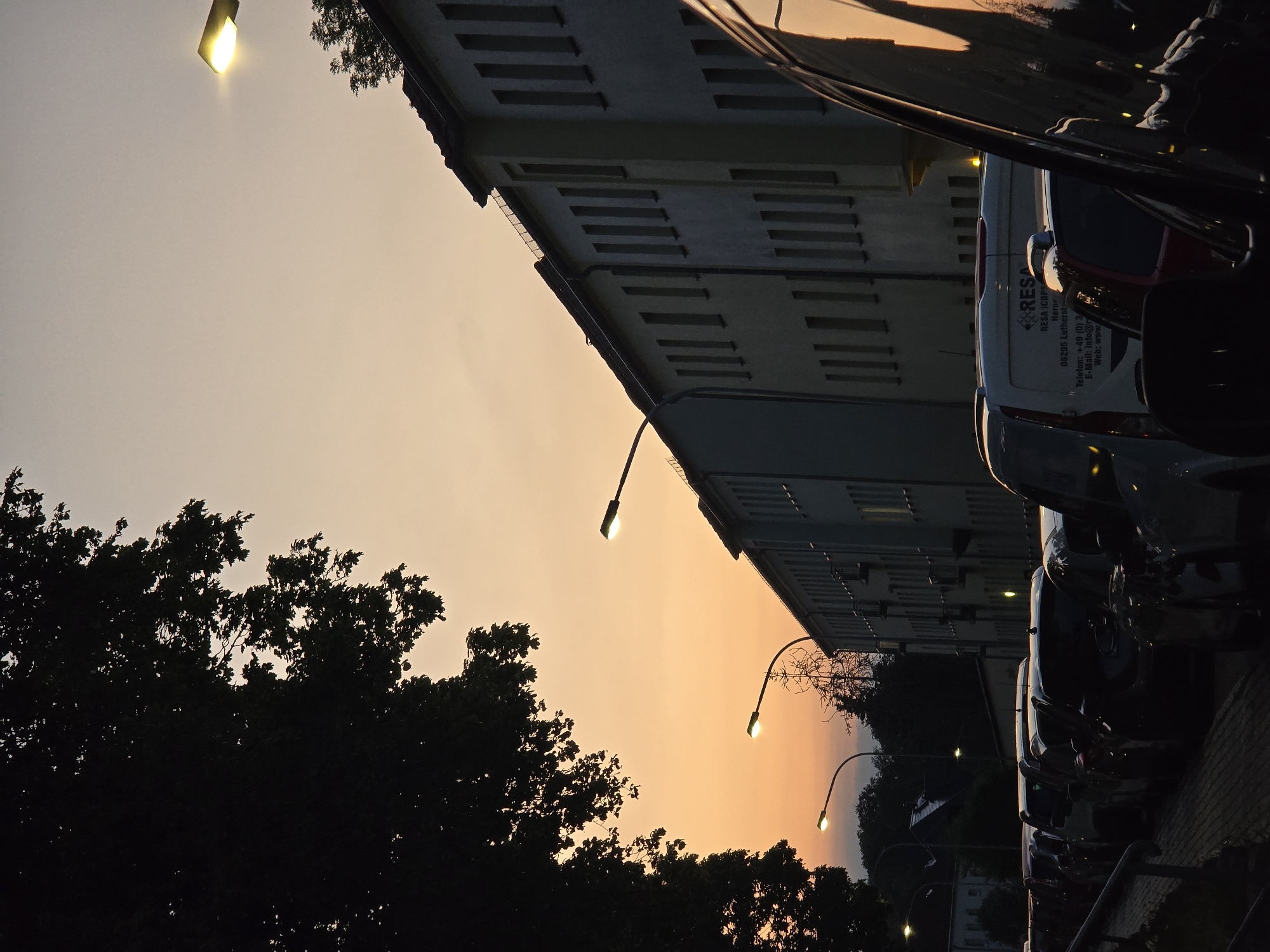 Here is a description of the image:
The image depicts a street scene during sunset, likely in Lutherstadt Eisleben, Germany, given the visible text "Lutherst" on a van. Several cars and a white RESA-branded van are parked on a paved area, adjacent to a multi-story building with repetitive rectangular window patterns. The building's facade is illuminated by the sunset, creating a warm hue. Street lamps line the street, their lights contrasting with the darkening sky. Silhouetted trees frame the lower left of the image. The sky displays a gradient of colors from pale yellow to deep orange, suggesting either sunrise or sunset.