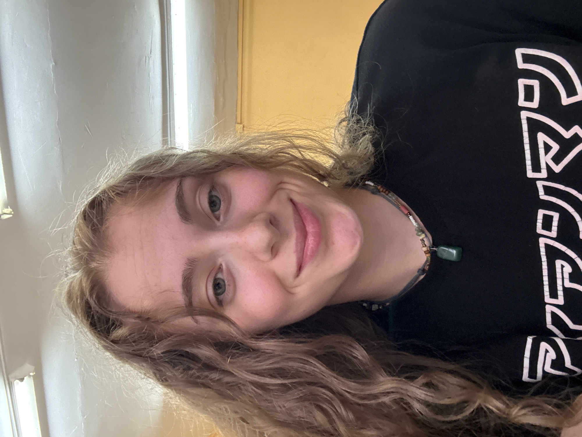 A young woman with long, wavy blonde hair is looking directly at the camera, smiling. She is wearing a black t-shirt with a necklace featuring a green pendant and various beads. The background appears to be indoors, with a yellow wall visible on the right and fluorescent lighting fixtures on the ceiling. The overall lighting is bright. There are no identifying landmarks or specific details that would indicate the location as Sofia, Bulgaria.