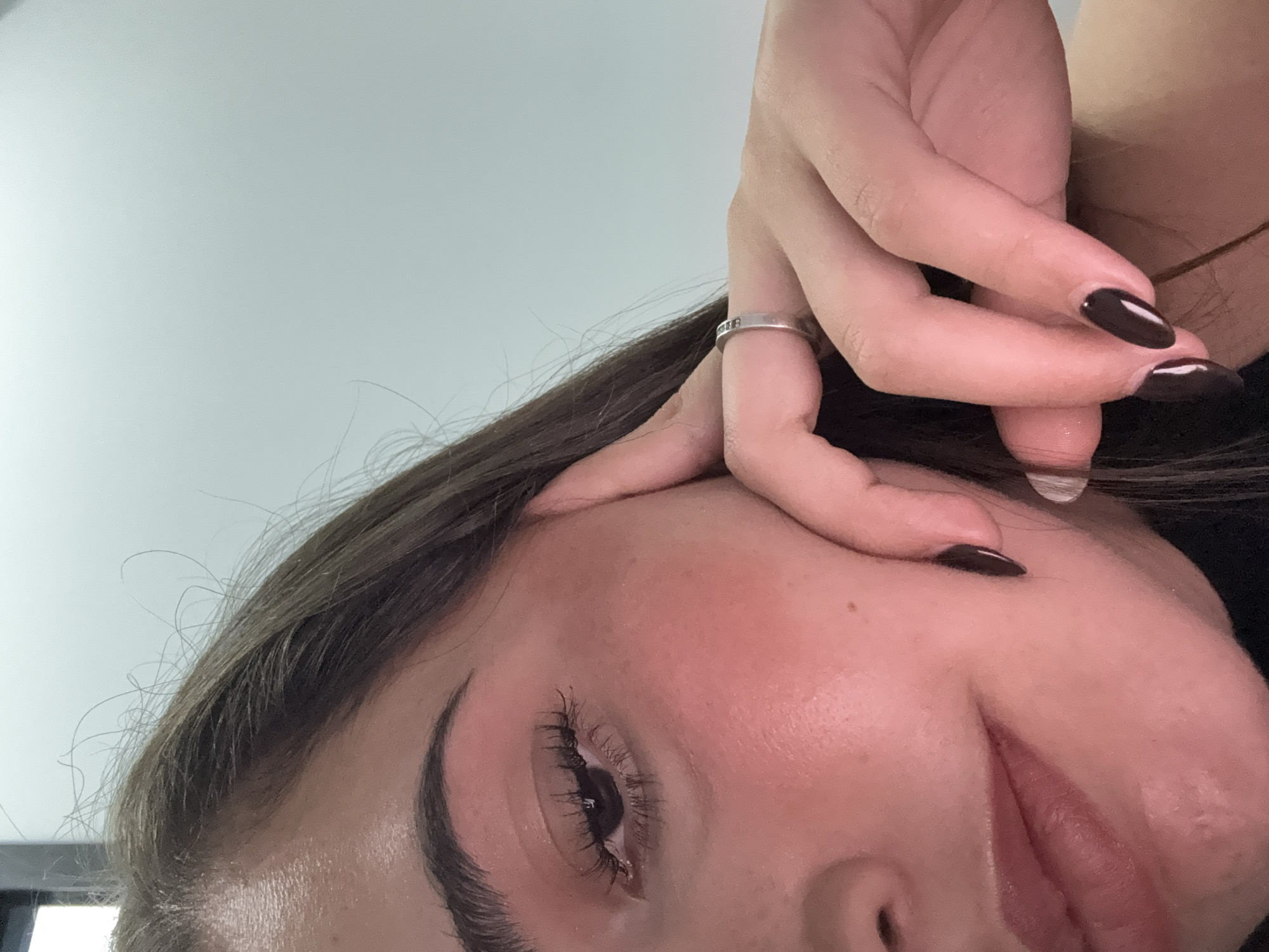 The image is a close-up, upside-down view of a person's face, with one hand partially obscuring the forehead. The person has dark eyebrows, eyelashes, and brown hair. The fingernails are painted a dark color. A ring is visible on one of the fingers. The background appears to be a plain, light-colored wall or ceiling.
There is insufficient information in the image to determine the location in the city of San Bernardino Tlaxcalancingo, Mexico.