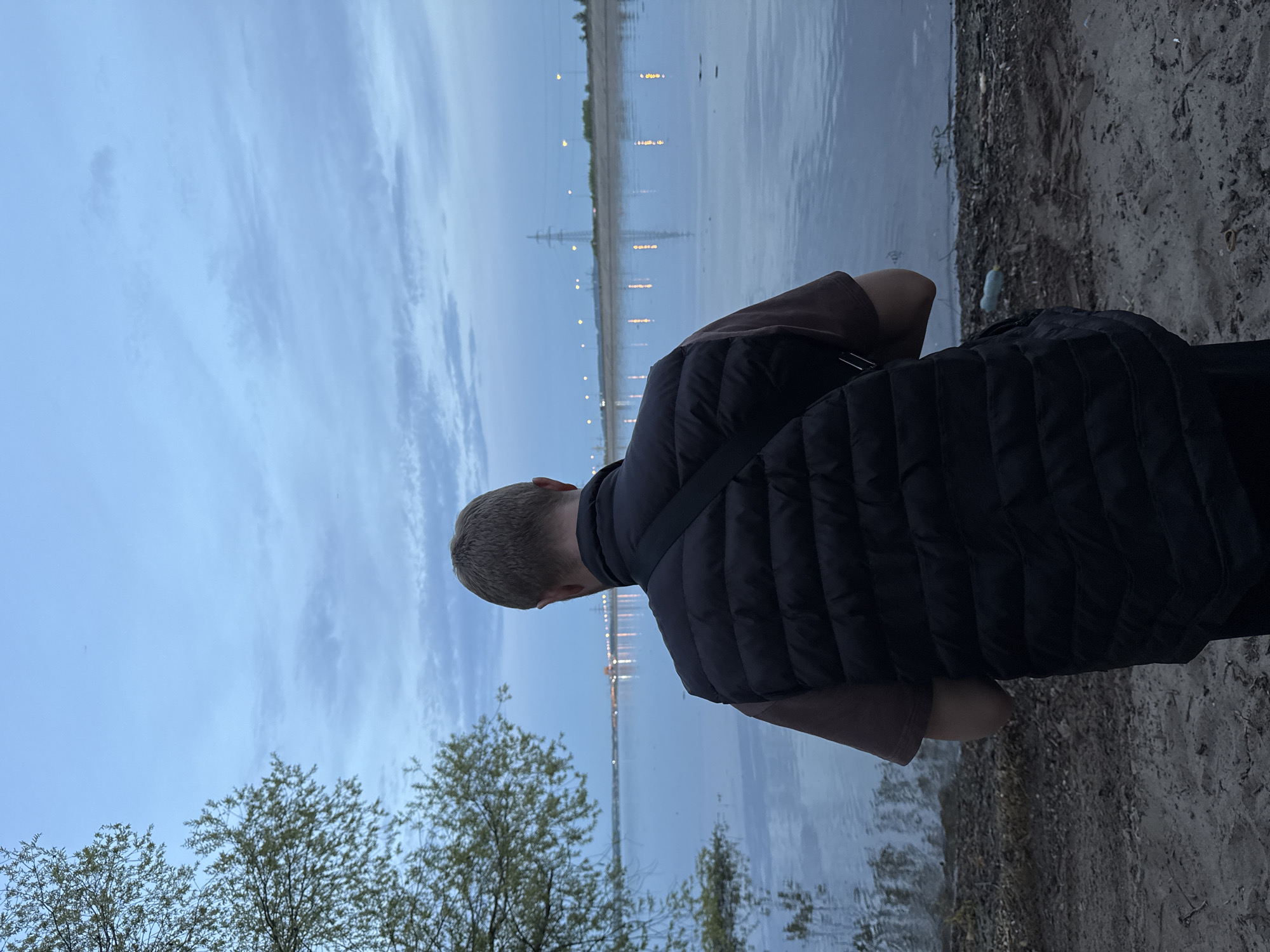 A photograph shows a person from behind, standing on the edge of a body of water. The person has short blond hair, wears a brown shirt, and a black puffer vest with a strap across their back. They are looking out at the water.
Across the water are lights, likely from buildings and streetlights, indicating an urban area. The reflection of these lights is visible in the water. Power lines and towers are visible on the other side of the body of water.
The ground on which the person stands appears to be sandy or gravelly, with sparse vegetation. Some trees are on the left side of the image. The sky is overcast with light blue and gray tones.
Based on the description and the lights across the water, this scene appears to be in Noviy, Russia.