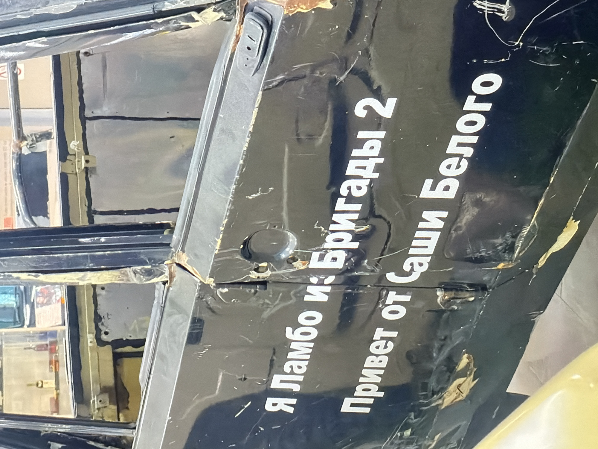 The image shows a black car door with Russian text written in white. The text reads: "Я Ламбо из Бригады 2 Привет от Саши Белого" (I'm Lambo from Brigade 2 Greetings from Sasha Belogo). The door appears to be damaged or in a state of disrepair, with peeled and ripped areas. The location and city are unknown.