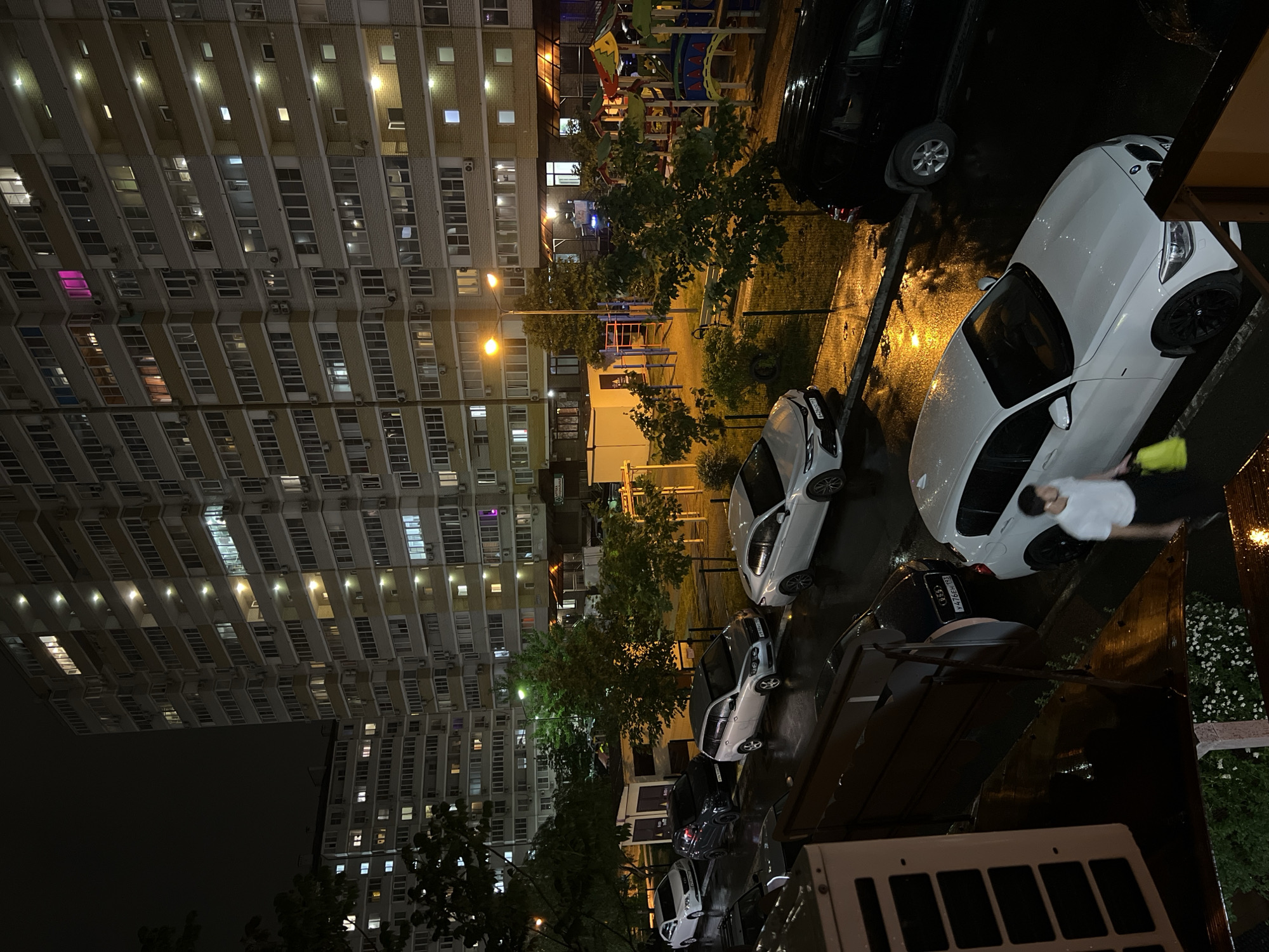 The image depicts a nighttime urban scene in Krasnodar, Russia. A tall residential building dominates the left side of the frame, its facade featuring rows of windows with lights emanating from some. In front of the building, there's a playground with visible play structures.
The right side of the frame contains a street or parking area with several parked cars, including a white BMW with the plate V416ER193 and a black car parked behind it. A man wearing a white shirt and dark pants is standing near the white car. The ground appears wet, possibly from rain. The lighting comes from streetlights and the illuminated windows of the building. Some greenery, likely trees and bushes, can be seen interspersed between the buildings and the street.