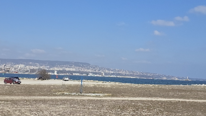 The image presents a panoramic coastal scene in Varna, Bulgaria, under a clear blue sky with scattered faint clouds. The foreground consists of an unpaved, barren expanse of light brown and gray earth mixed with sparse, dry vegetation. A single, upright blue pole is situated in the mid-right foreground.
Immediately beyond the foreground, the dark blue waters of the Black Sea extend horizontally, bordered by a low-lying rocky or concrete breakwater. Three vehicles are visible on the unpaved terrain near the water's edge: a burgundy SUV and a dark-colored sedan are parked on the left, and a lighter-colored sedan is positioned further to the right. A small, bare deciduous tree stands centrally in the midground, close to the water. Within the water, a green conical buoy is visible on the left, and a red cylindrical buoy is located further to the right, marking navigational points.
The background features the extensive urban landscape of Varna city, characterized by numerous multi-story buildings spreading along the coastline and ascending into hills beyond. The city's port area contains several docked vessels and yachts. A prominent white lighthouse or navigational tower is visible on a distant peninsula to the far right, while another white tower-like structure is discernible on the distant shore line, to the right of the central tree. No individuals or specific activities are identifiable in the scene.