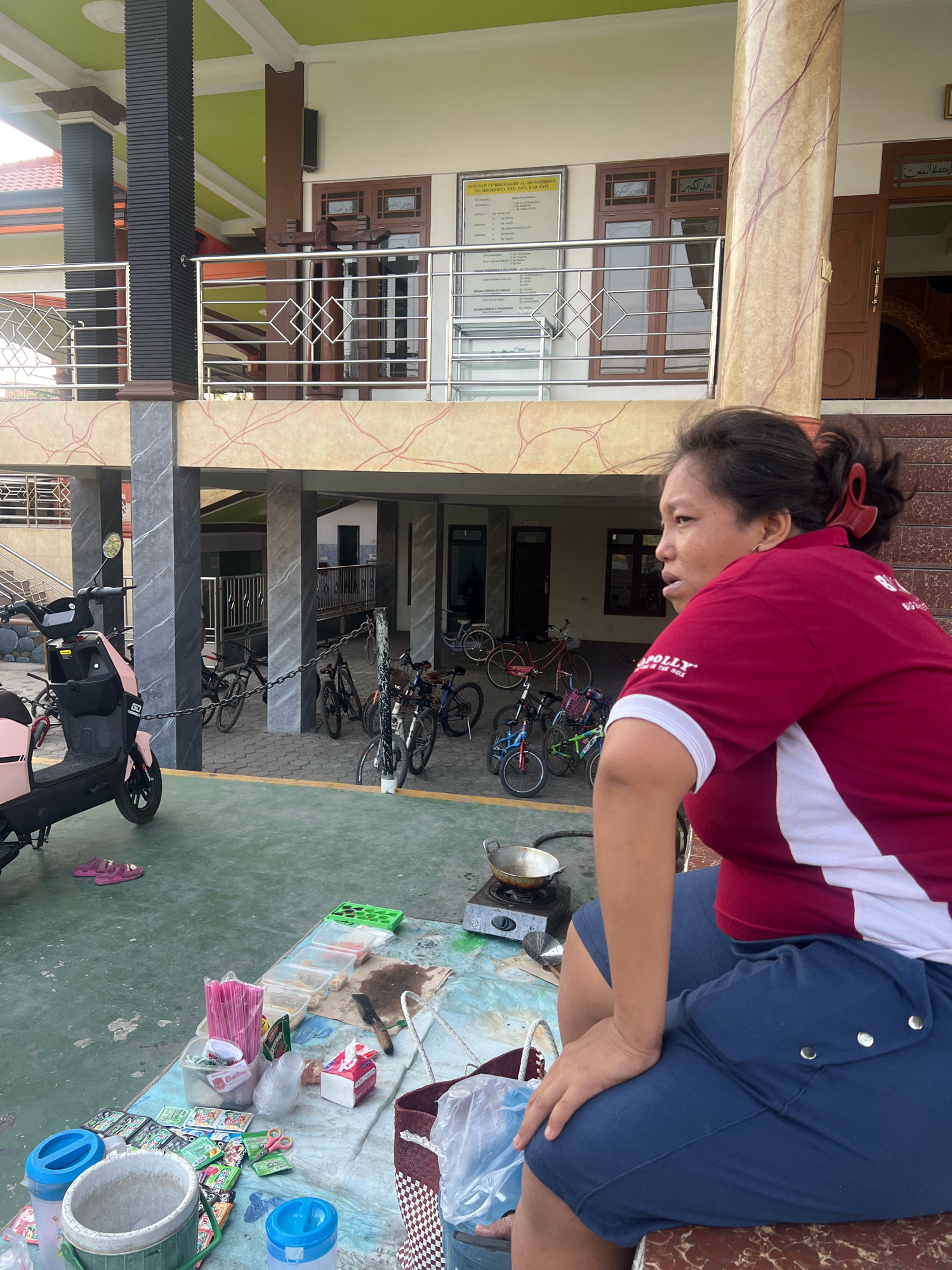 A medium shot captures a woman sitting on a low bench, facing right, in an outdoor area. She has dark hair pulled back in a ponytail with a red hair tie. She is wearing a red polo shirt with white trim and navy blue shorts. In front of her, a table is laid out with various items, including packets of food, drinking straws, cups, tissues, and a small gas stove with a pan on top. Behind her, several bicycles are parked, and a pink electric scooter is visible to the left. The background reveals a building with a green ceiling and patterned columns, suggesting a public space or an institution. The scene appears to be in Kecamatan Pati, Indonesia, given the presence of typical Indonesian street food stall items and the architectural style.