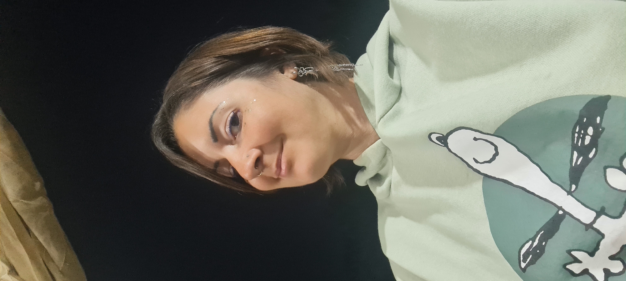 A medium shot of a woman, angled slightly to the left. She has short brown hair, fair skin, and a nose ring. There is what appears to be glitter or light reflecting near her left eye. She is wearing a light green hoodie with a graphic design of what looks like a cartoon character holding crossed objects, possibly bones or sticks. The background is a dark, uniform black, with a blurry tan object at the left edge. The image appears to be taken indoors or in a setting with controlled lighting. The location is reported to be in Lachish Regional Council, Israel.