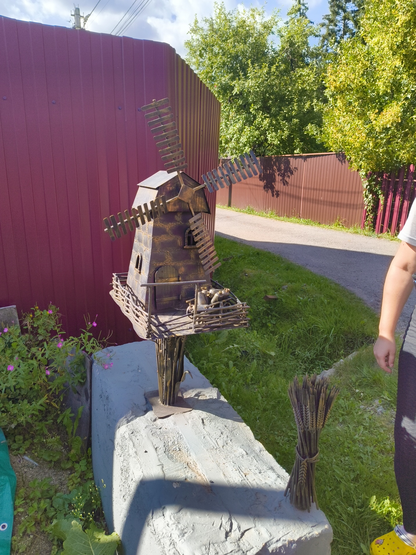 The image depicts a decorative metal windmill placed on a concrete structure in an outdoor setting. The windmill is brown and appears to be aged. It has four prominent blades and a small balcony area with a rustic fence. Inside the fenced area, there are several small, metallic objects that resemble bottles or jugs. A bunch of metallic wheat stalks is tied with twine and placed on the concrete structure to the right of the windmill. To the left of the windmill, there are some green plants with small pink flowers and a green tarp visible. In the background, there is a tall, maroon corrugated metal fence, a pathway, and lush green trees under a cloudy blue sky. A person's arm and legs, wearing a grey shirt and black pants with yellow Crocs, are visible on the right side of the image, suggesting their presence near the scene. The overall environment appears to be a garden or yard area. The location is specified as SNT Pravdist-2, Russia.