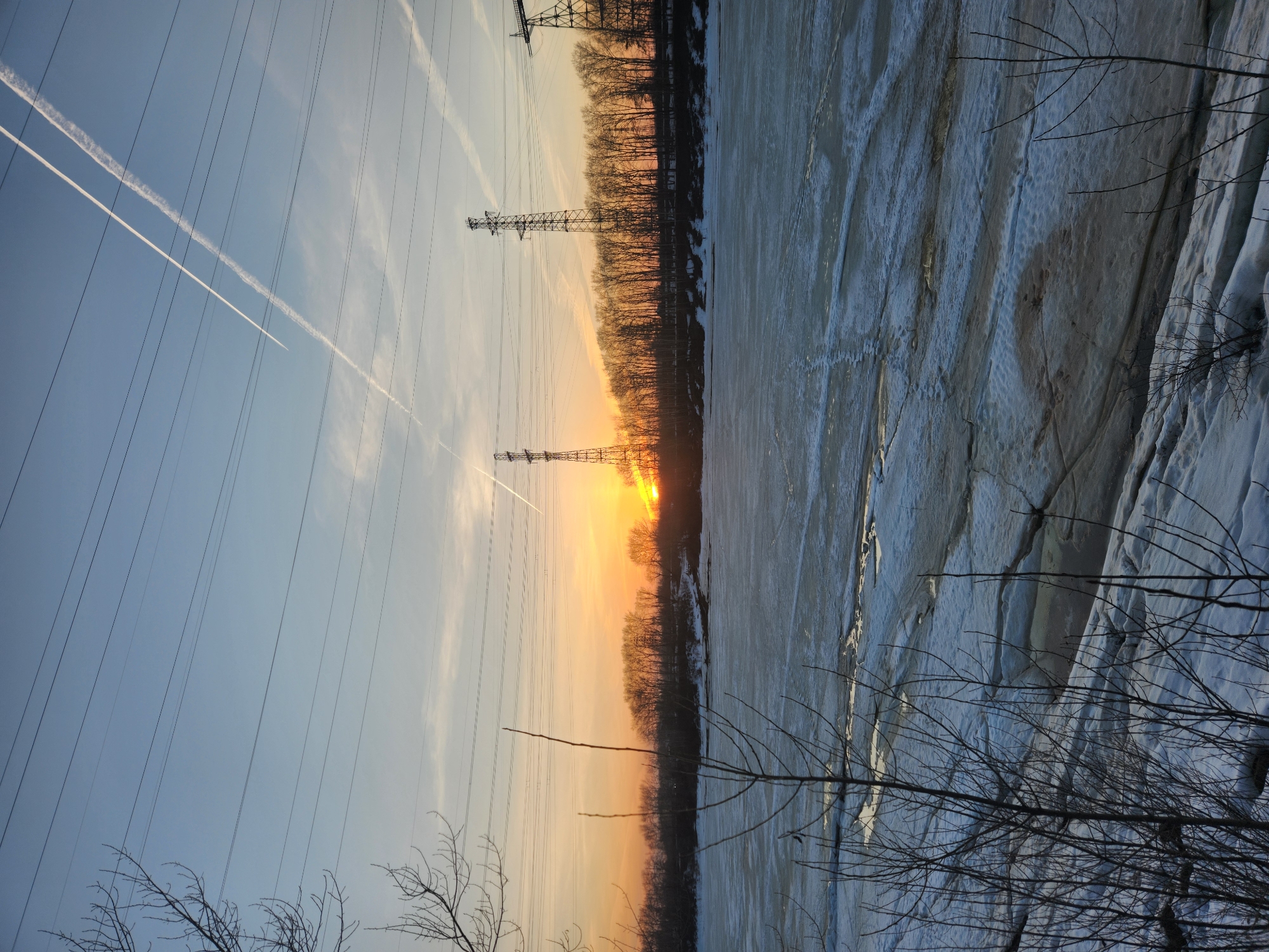 The image depicts a winter scene in Chaikovsky, Russia. The sun is setting, creating a gradient of orange to blue in the sky. Power lines and electrical towers are prominent in the frame. The foreground shows a snow and ice-covered river or body of water. Bare trees and branches are visible along the riverbank and in the lower part of the frame. Contrails from airplanes streak across the sky.
