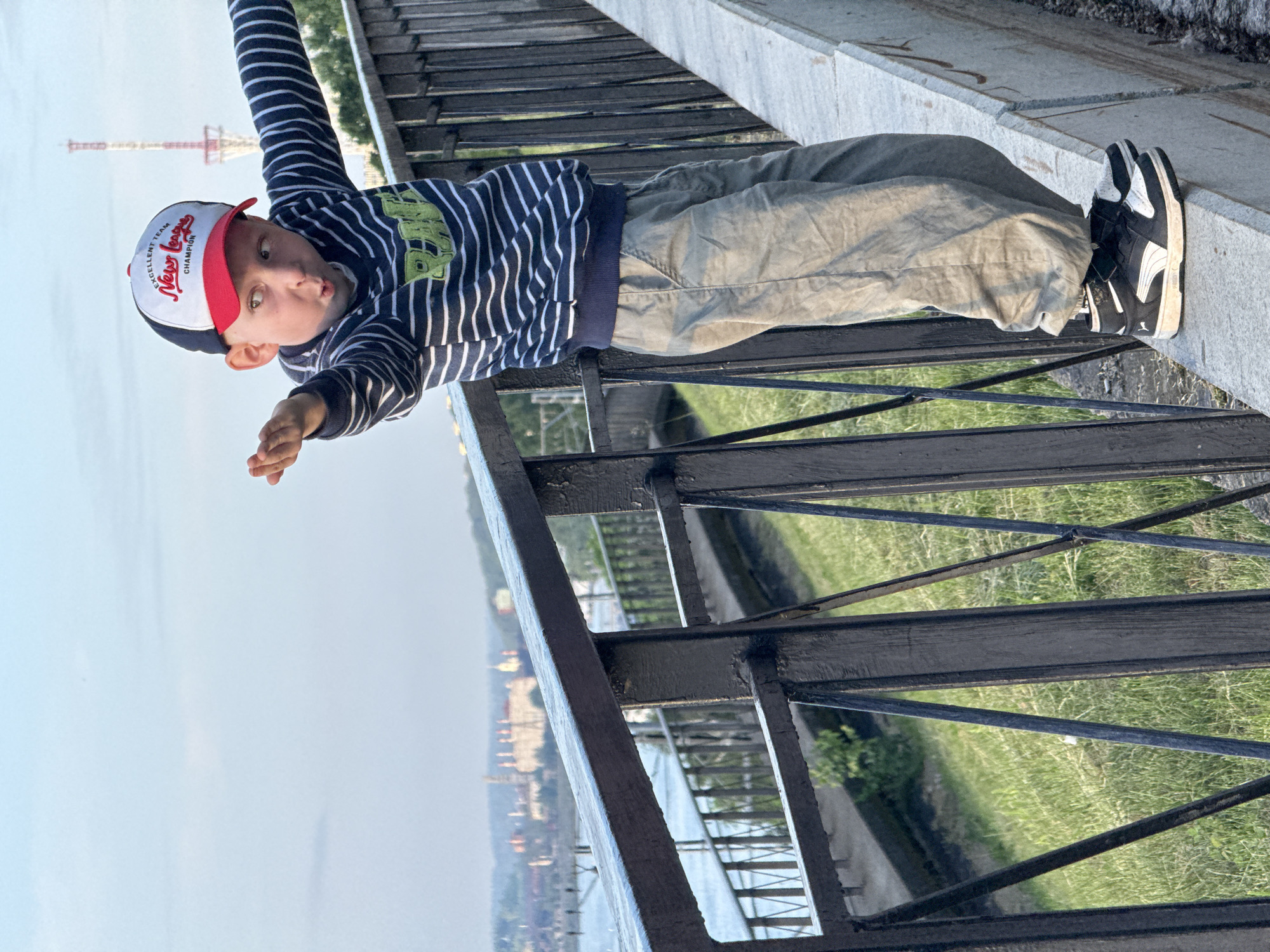 A young boy wearing a striped sweater, khaki pants, and a baseball cap stands on a concrete ledge next to a black metal fence. He is looking to his left with his mouth open in a surprised expression and his arms outstretched as if to balance himself. In the background, a body of water is visible, and beyond that, a city skyline with several buildings and a tall communication tower. The setting appears to be a park or embankment overlooking a city. The boy's shoes are black and white Puma sneakers. The boy is positioned on a railing or ledge, indicating a potentially precarious balancing act. The city in the background, with its distinctive architecture and communication tower, is characteristic of Perm, Russia, possibly taken from the embankment near the Kama River.