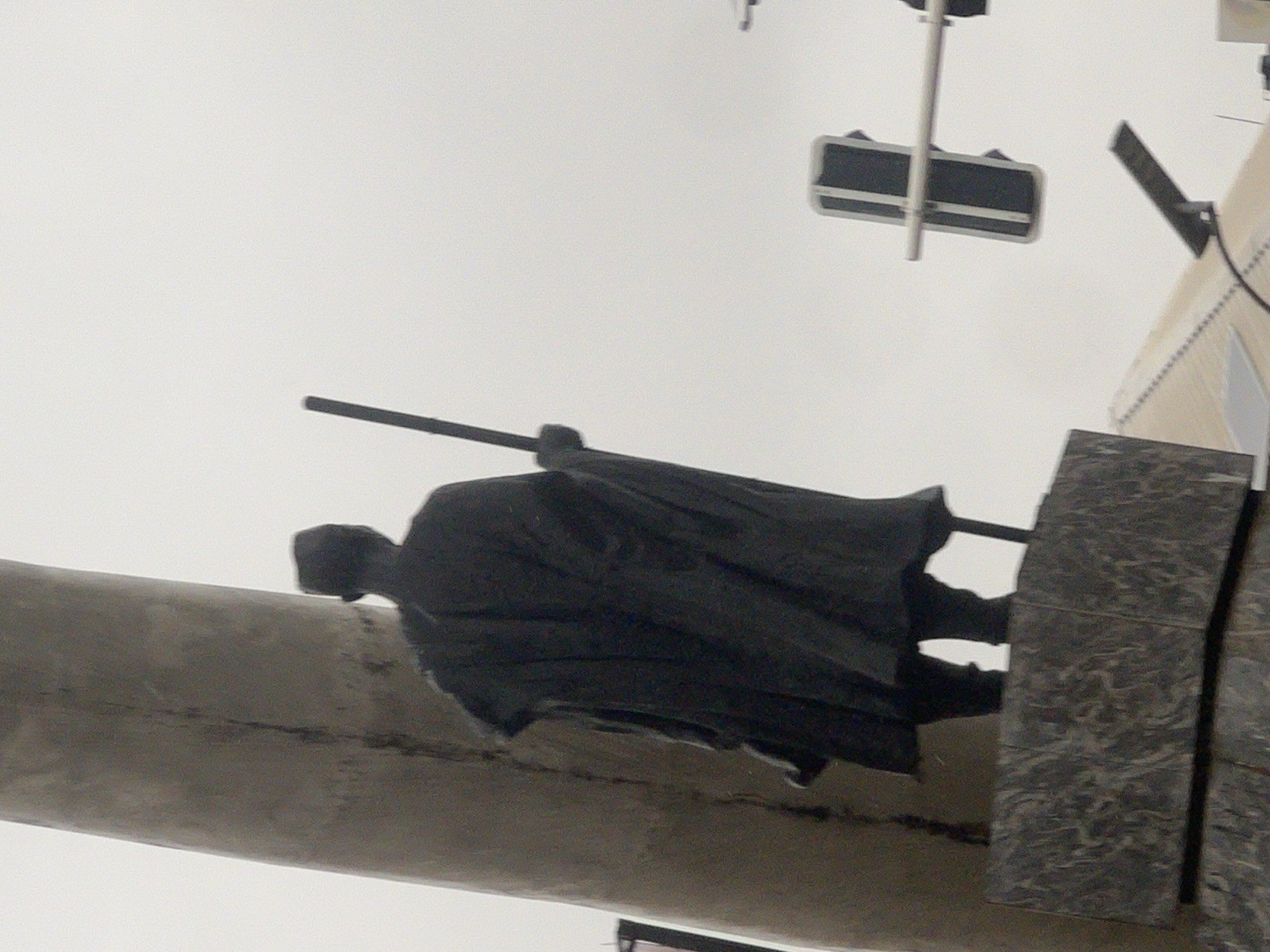 The image shows a statue of a man holding a stick, located in Onitsha, Nigeria. The statue is dark and appears to be made of stone. It is standing on a stone pedestal. In the background, there is a traffic light and a cloudy sky. The statue is positioned to the left of the frame, and the traffic light is on the right. The overall scene is somber, with the grey sky and dark statue contributing to a somewhat muted atmosphere. The statue's stance suggests a figure of authority or contemplation.