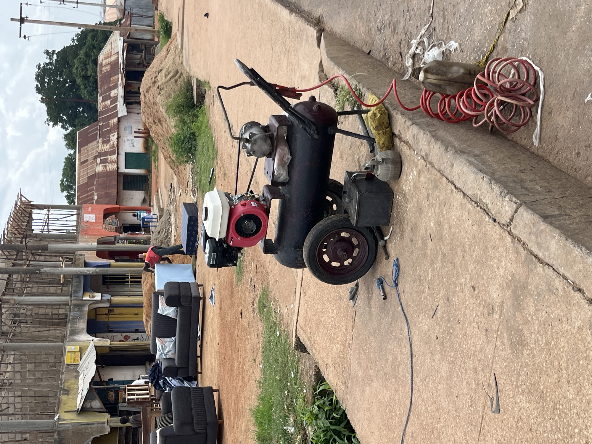 The image depicts an outdoor scene in Bukuru, Nigeria, likely near a commercial area or construction site. In the foreground, a large, black air compressor with a red and white engine is positioned on a concrete surface. Various tools and accessories, including a coiled red hose, a dark metal container, and a bottle, are scattered around the compressor. To the left of the compressor, two dark grey sofas with patterned cushions are placed on the reddish-brown earth.
In the midground, several buildings with corrugated metal roofs and plastered walls are visible. Some buildings appear to be under construction, with exposed wooden and metal framing. A pile of sand is situated next to one of the structures. A person in a red shirt is seen interacting with a large blue object near the sand pile.
Further in the background, more buildings and trees are present, suggesting a semi-urban or developing area. The overall environment appears dusty and unpaved, consistent with a scene in Bukuru, Nigeria. The sky is overcast with visible power lines and poles.