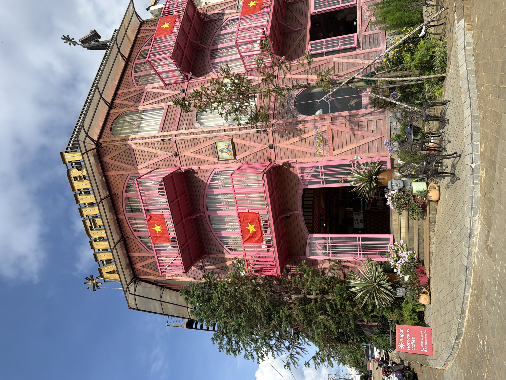 The image captures the facade of a uniquely designed building in Măng Đen, Vietnam. The building's exterior features a pink wooden structure with rounded balconies, each adorned with a Vietnamese flag. There are oval-shaped windows, and a distinctive sign on the building's left side. A variety of plants and flower pots are arranged along the entrance steps, adding to the building's whimsical charm. The building faces a brick-paved sidewalk. A sign for "Auguri Homestay Coffee" is visible near the base of the building. The sky is partly cloudy, and the building's architectural elements create a visually arresting and unusual scene.