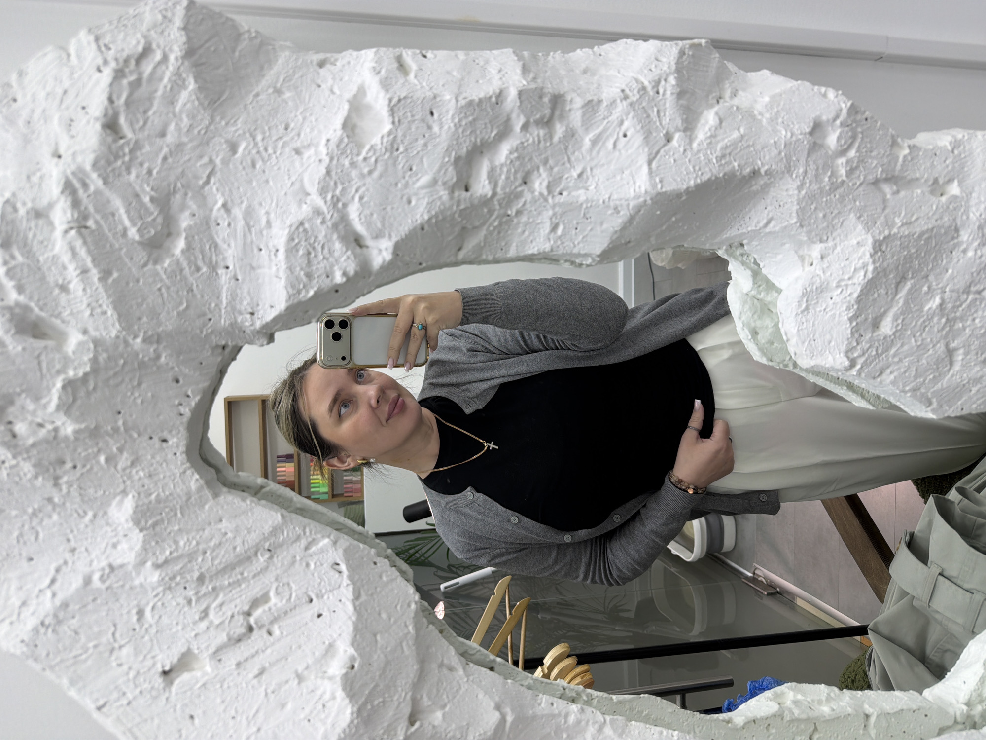 A young woman takes a mirror selfie, her reflection framed by a distinctive, irregularly shaped white border. The border appears to be made of a heavily textured material resembling rough plaster or a fractured, organic form, giving the mirror a sculptural quality.
The woman, positioned from the chest up, holds a light-colored smartphone, possibly an iPhone, with multiple camera lenses, capturing her image. She has light blue eyes and her hair, with lighter highlights, is pulled back. She wears a black, short-sleeved top, a grey button-up cardigan, and light-colored trousers. A delicate gold-colored necklace with a small cross pendant hangs around her neck, and bracelets adorn her right wrist. A ring is visible on her left hand, which holds the phone, while her right hand lightly touches her midsection. Her gaze is directed towards the camera of her phone, and her expression is neutral.
The background visible in the mirror reflection indicates an indoor setting, likely a studio or a contemporary space. To the left, behind her head, a wooden shelving unit filled with various colorful items, possibly art supplies or product samples, can be seen. Other faint reflections hint at more furniture or display items. The lighting is bright and diffused, suggesting daytime indoors. There is no visible text in the image. The unique, artistic mirror frame is a prominent feature of the scene. The image was captured in Moscow, Russia.