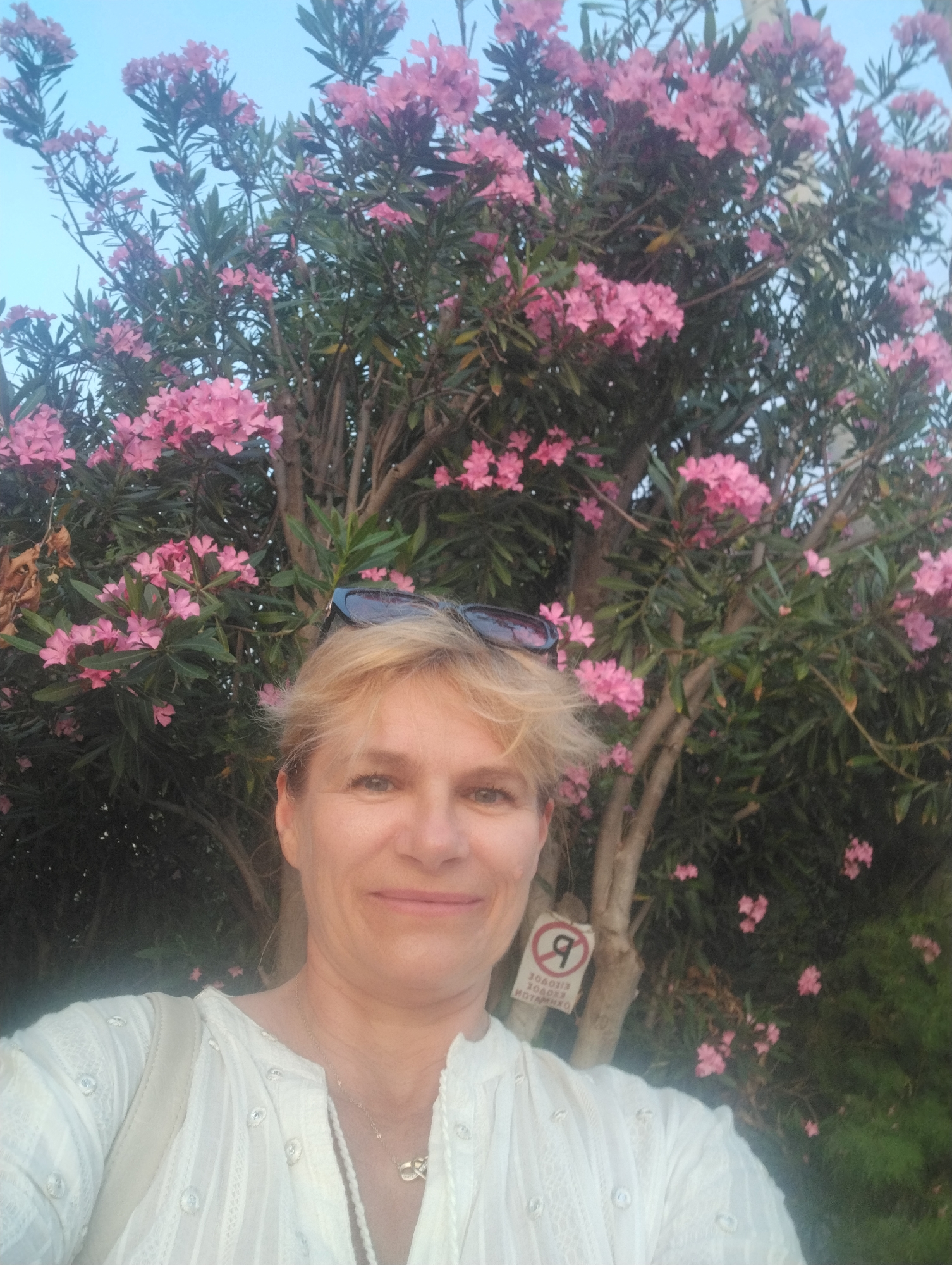 A medium shot captures a woman with blonde hair posing for a selfie in front of a tree laden with pink flowers. The woman is looking at the camera and smiling gently. She is wearing a white, embroidered shirt with a delicate necklace. Sunglasses are perched on her head. Behind her, the tree's branches are covered in numerous pink blossoms, with lush green leaves providing a vibrant backdrop. The sky is a clear blue, suggesting it is daytime. A small, circular sign with a red border and a white symbol inside is visible on the tree trunk to the woman's right. The symbol appears to be a stylized "R" or similar character, with some illegible text below it. The overall scene evokes a feeling of warmth and natural beauty, possibly during a spring or summer day in an outdoor setting.