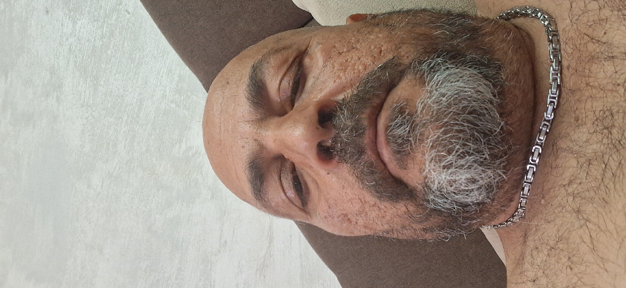 The image displays a close-up, low-angle portrait of an adult male, observed from a slightly upward perspective. The man is bald on the crown of his head and possesses a full beard and mustache, which are predominantly gray, particularly around the chin, with darker hairs mixed throughout. His eyes are partially closed or squinting, and his eyebrows are dark. The skin on his face appears textured. Around his neck, a silver-colored chain with distinct rectangular or square links is visible, resting on his upper chest, which is covered in dark hair.
The background behind the man consists of two elements: a light grey or off-white wall or textured surface with subtle horizontal and diagonal patterns occupies the upper left and central top portions of the frame. To the right and behind the man's head, a section of plain, dark brown upholstered fabric, consistent with a couch or pillow, is visible. The lighting is soft and even.
There are no identifiable landmarks, architectural features, or other contextual clues within the image to determine a specific geographical location, including the city of Simeonovgrad, Bulgaria.