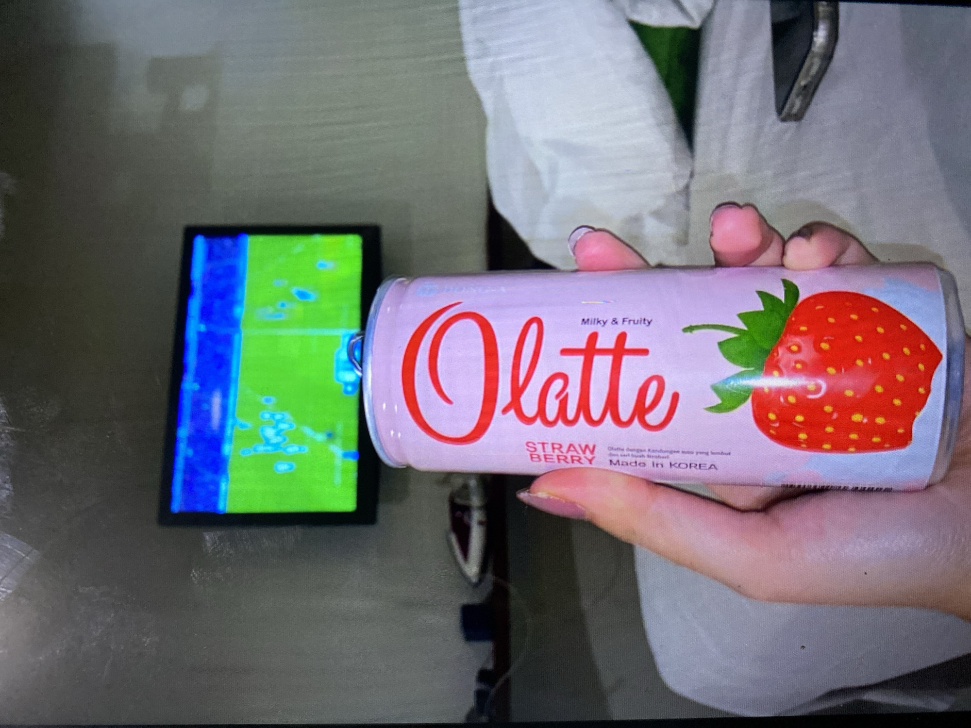 The image shows a close-up of a person's hand holding a pink aluminum can with a strawberry design and "Olatte" written in large red script. The can also has text indicating "STRAW BERRY" and "Milky & Fruity." In the background, a television screen displays a soccer game. The environment appears to be a room, possibly a bedroom, with a white duvet and a shelf with some items on it. The presence of a television and indoor setting suggests this could be a domestic environment. The overall scene is casual. The location of Kalitinggar, Indonesia, is not visible in the image.