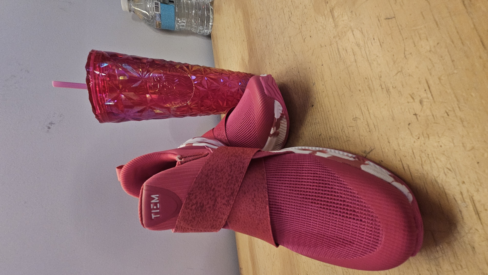 The image shows a pink drinking cup with a straw, a pair of pink TIEM shoes, and a plastic bottle of water. The items are on a light brown surface. The background is a white wall.
There is no indication that the image was taken in the city of Puebla, Mexico.