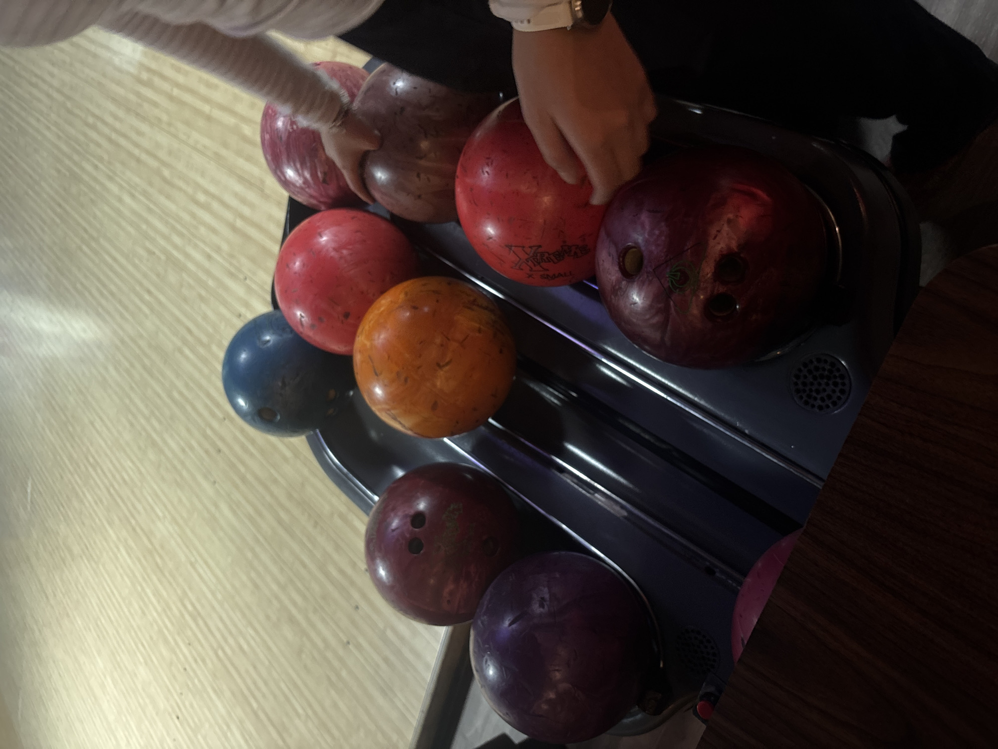 A high-angle, close-up shot captures a bowling alley scene in Naucalpan de Juárez, Mexico, focusing on a collection of bowling balls in a return rack. A person's arm, partially covered by a white sleeve, and hand are visible at the top-center of the frame, reaching towards the balls, suggesting the activity of selecting or returning a ball.
The dark grey or black rack holds numerous bowling balls of various colors, including bright red, orange, dark blue, and different shades of maroon and purple. The light-colored wooden floor of the bowling alley, featuring distinct parallel planks, occupies the left portion of the image. A dark wooden surface, possibly a table, is visible on the far right.
Notable details include text visible on one of the red bowling balls, which clearly reads "X-TREME" and "X SMALL," indicating its brand and size. Another dark purple/maroon bowling ball features a subtle, spider-like graphic near its finger holes. The indoor setting, illuminated by artificial light, does not provide cues for the time of day.