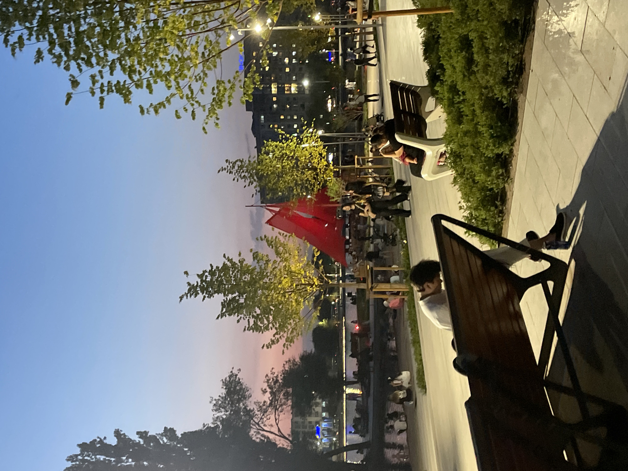 The image depicts an evening scene at the Sairan Lake in Almaty, Kazakhstan. A clear, pale blue sky transitions to a soft pink and orange hue near the horizon. The lake is in the mid-ground, with people sitting and walking along its edge. In the foreground, there are several park benches, one of which is occupied by a person leaning forward with their head down. Beyond the lake, apartment buildings are visible, with lights illuminating their windows. Trees with green foliage are scattered throughout the scene, some illuminated by streetlights. A large red fabric structure is visible in the mid-ground. The ground is paved with light-colored rectangular tiles, and there are patches of green bushes.