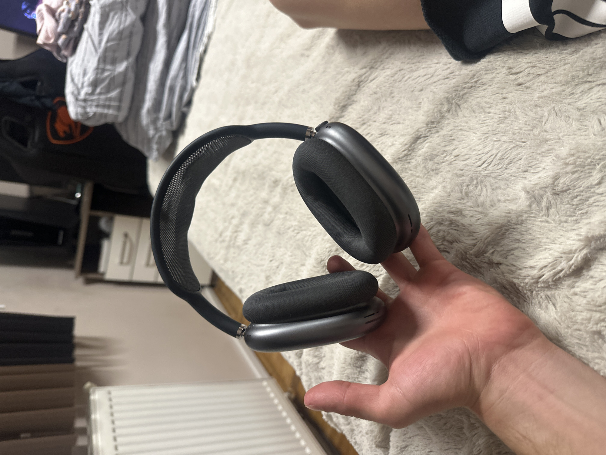 The scene is located in the city of Saraia, Bulgaria. The image presents an indoor setting, with a person's left hand prominently displaying a pair of Space Gray Apple AirPods Max headphones. The headphones feature a black mesh headband and black ear cushions, appearing to be in good condition. The hand and headphones rest upon a light-colored, textured, shaggy fabric surface.
In the background, a white panel radiator is visible on the left, adjacent to a light-colored wall. Above the radiator, brown and darker curtains are partially visible. To the right, a light-colored dresser or cabinet with two visible white drawers and silver pull handles is present, topped by a dark, rectangular object, possibly a monitor or television. Further right, a dark object with an orange, stylized cougar head logo is partially visible. A stack of folded textiles, including a pink garment, a striped gray item, and a plain gray item, is situated on what appears to be a bed. On the far right, a bare leg and a foot wearing a black and white sock or slipper are partially visible.